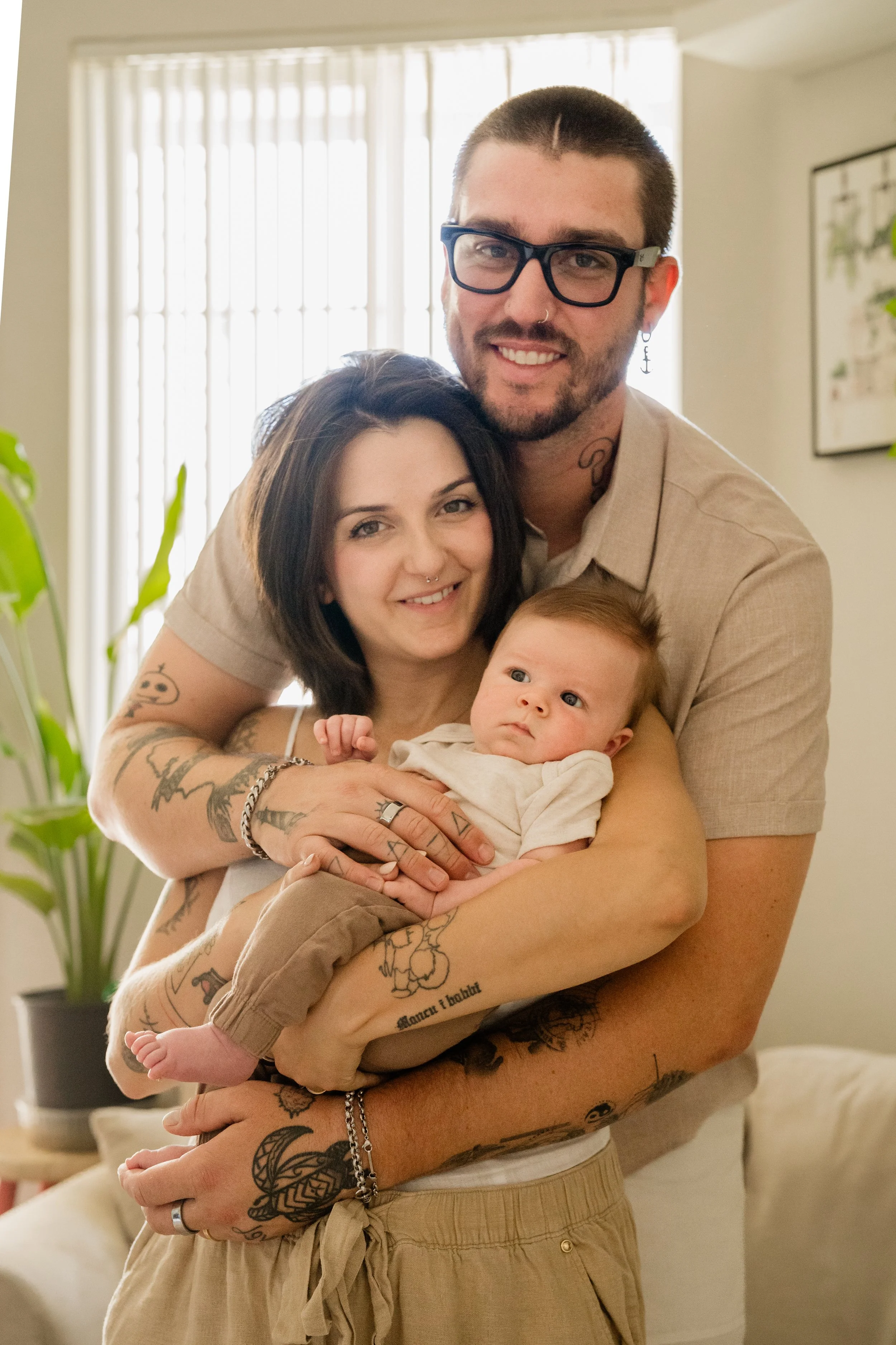 A father stands behind the mother with his arms wrapped around her and their baby, all three looking toward the camera in front of a bright living room window