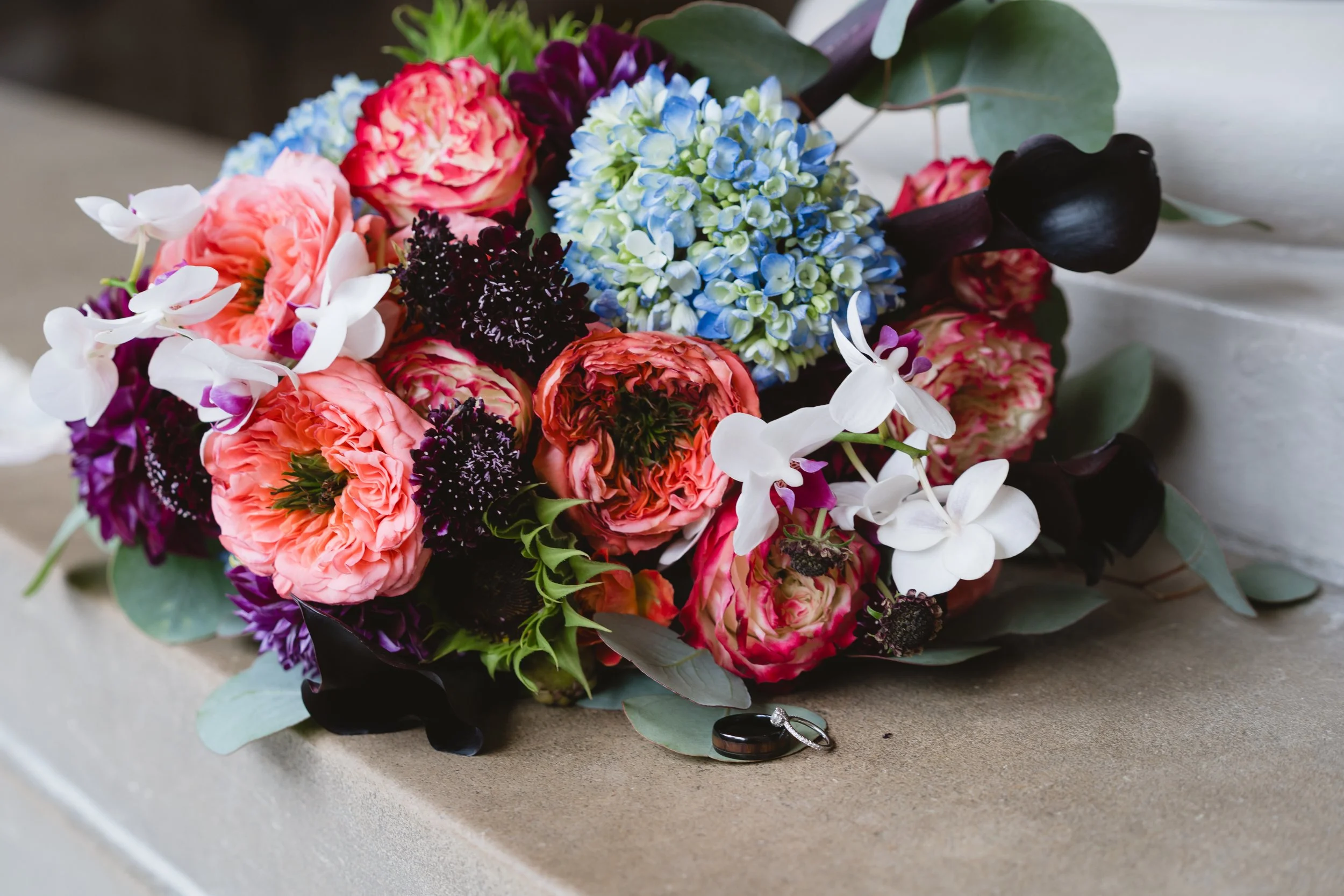 Bridal bouquet of coral peonies, purple dahlias, and blue hydrangea resting on a marble ledge beside the wedding rings.