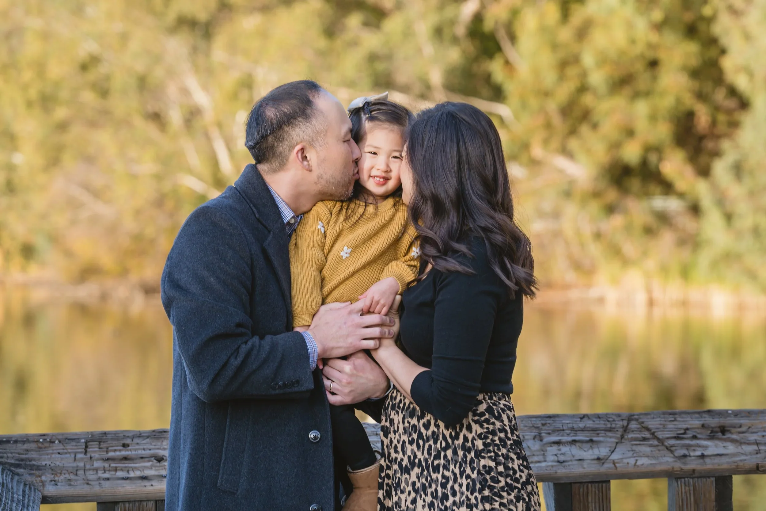 Mom and dad each kiss one of their daughter's cheeks at the same time while she smiles, framed close against a blurred golden lakeside backdrop.