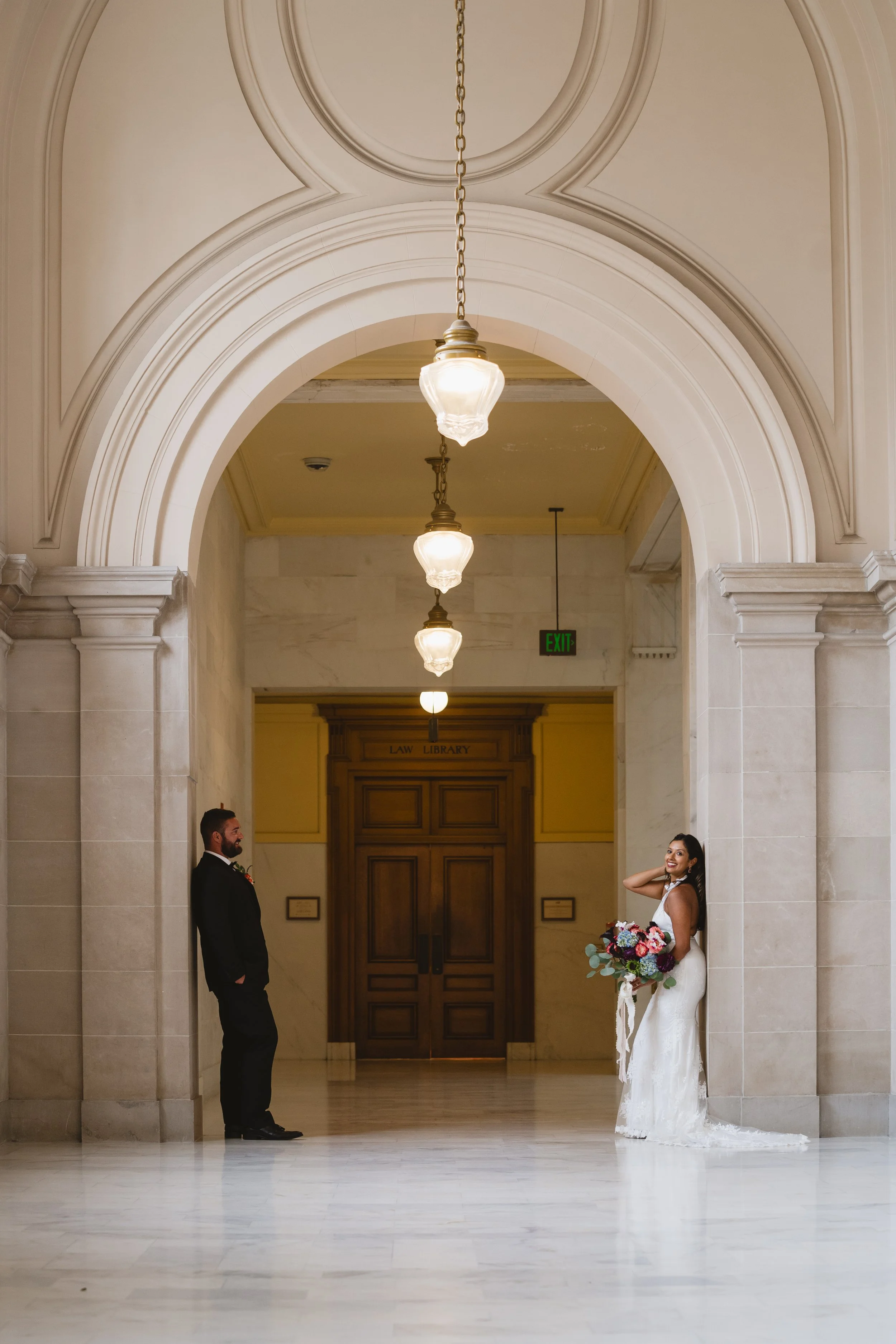 Bride and groom standing apart against columns in a marble corridor near the Law Library entrance, bride glancing back over her shoulder.