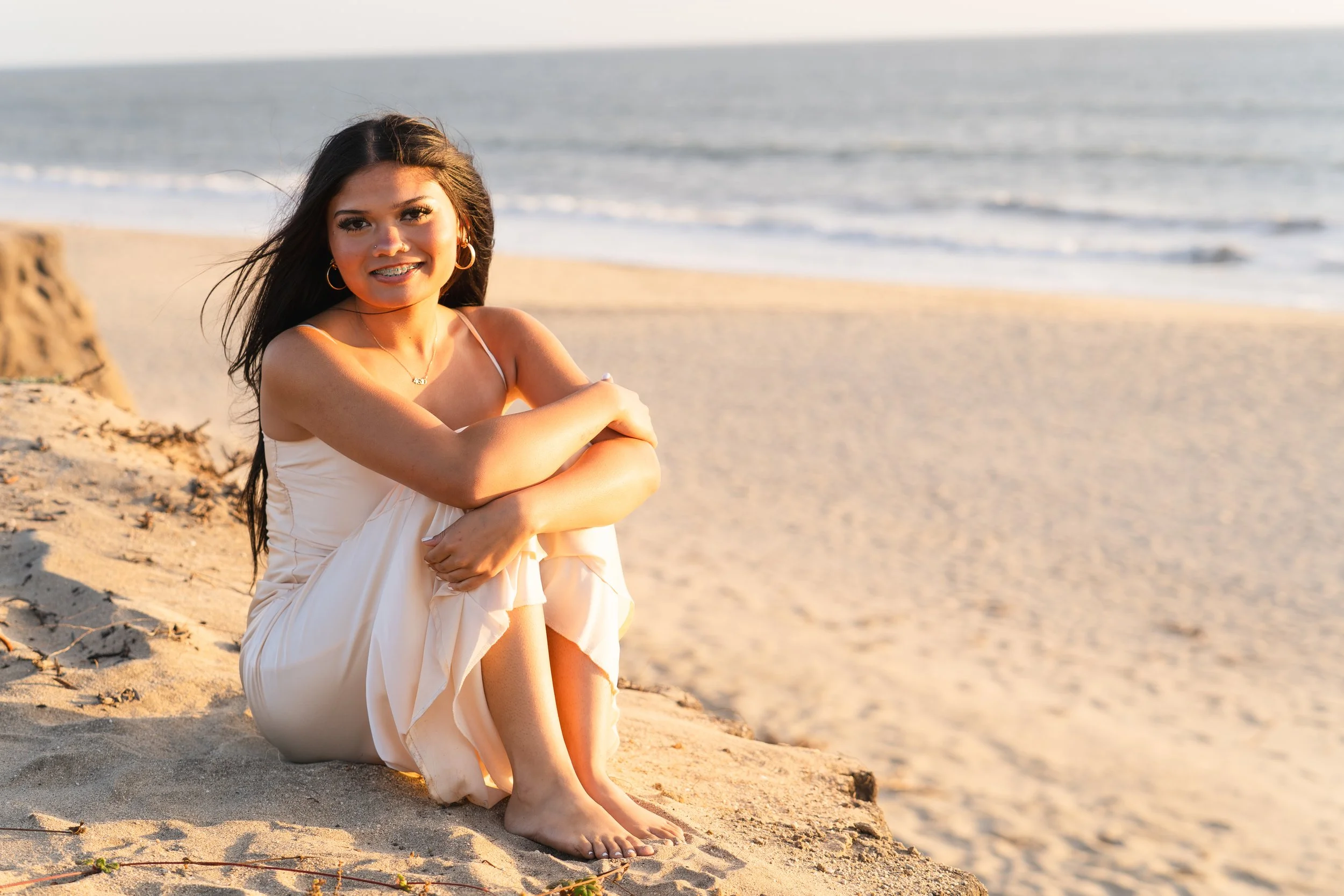Senior sitting in the sand with her arms around her knees, smiling at golden hour on a Half Moon Bay beach