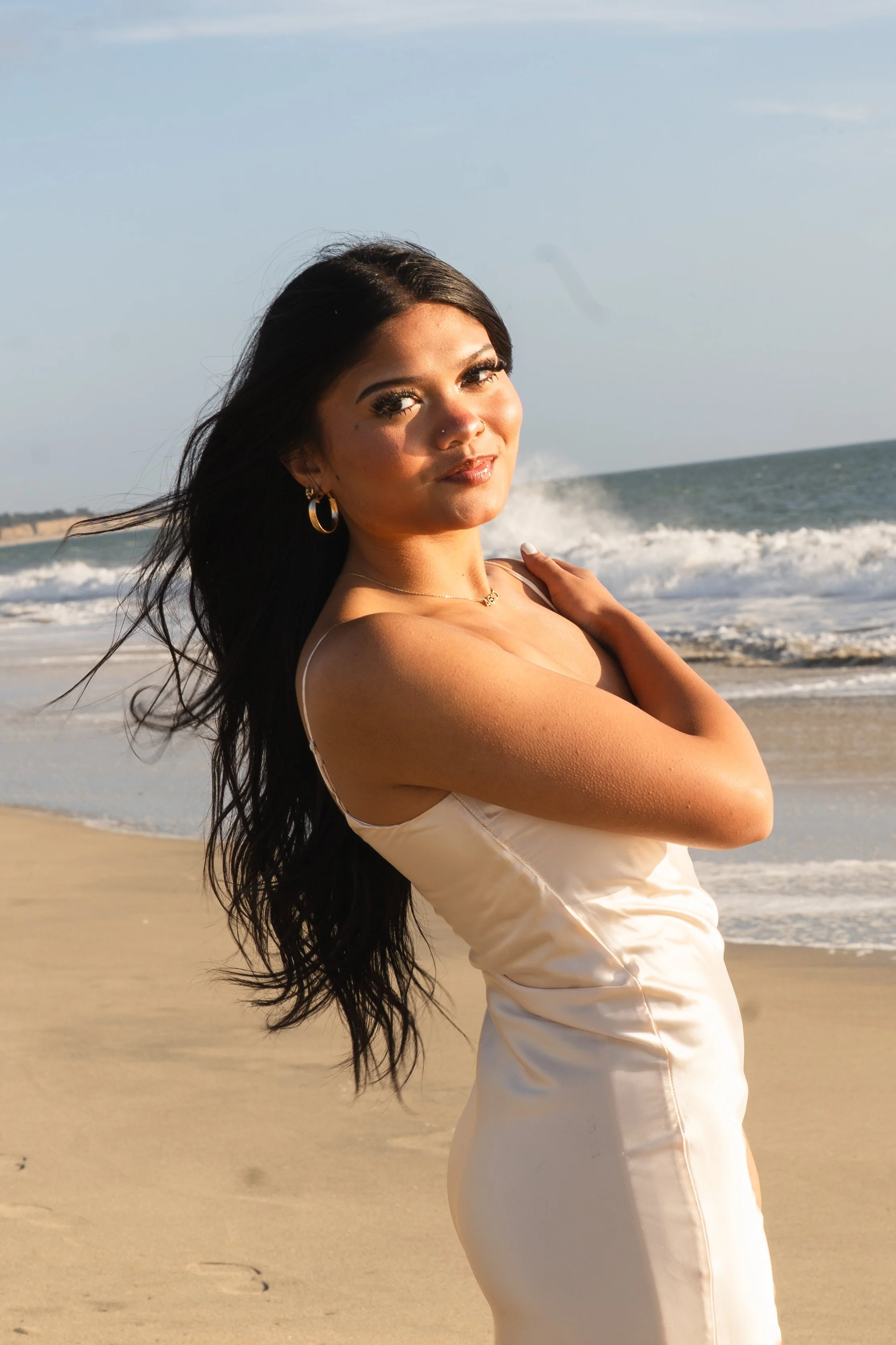 High school senior in a cream satin dress smiling over her shoulder as waves break behind her on a Half Moon Bay beach