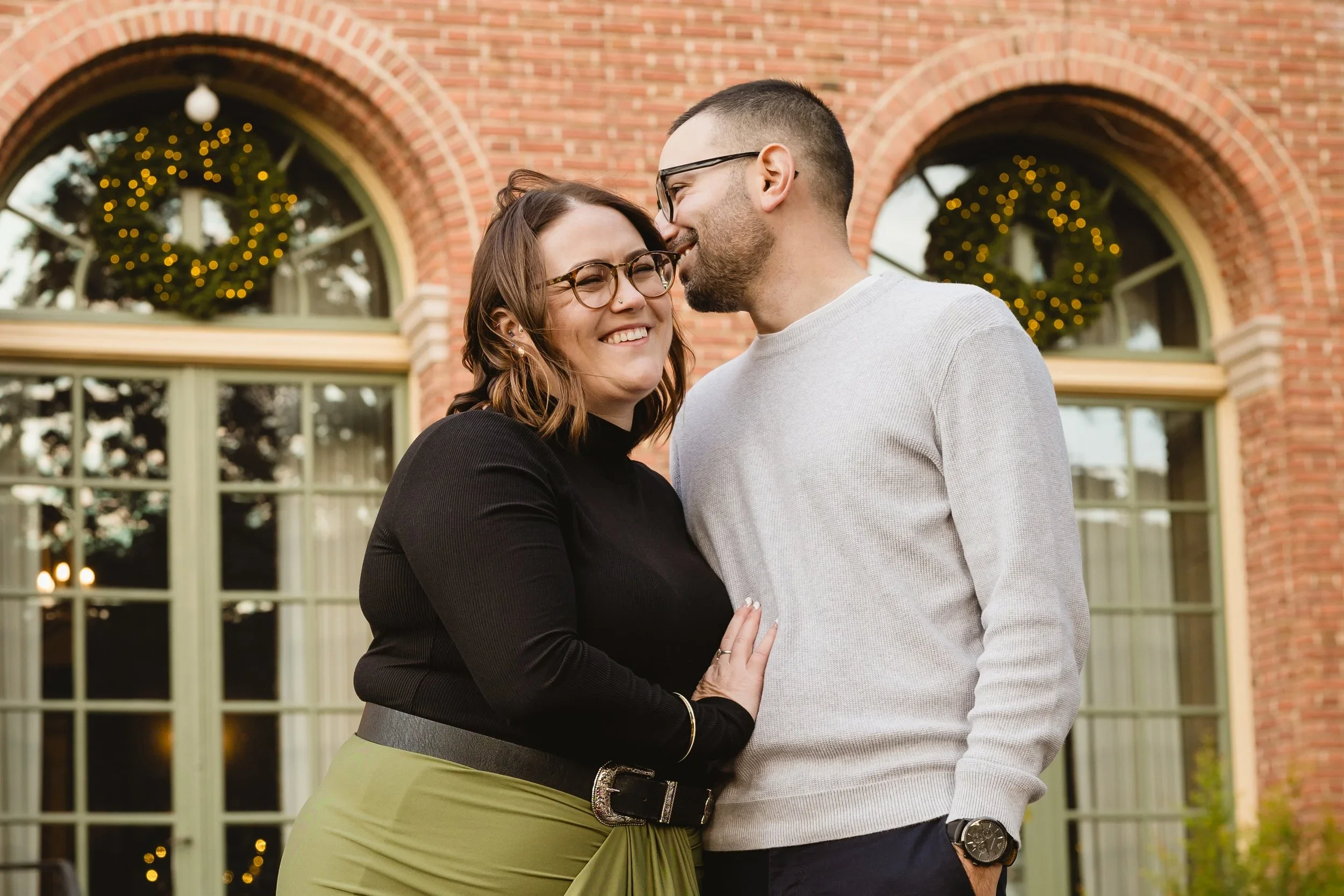 He leans in near her temple while she smiles toward the camera, standing under brick arches hung with lit greenery wreaths.