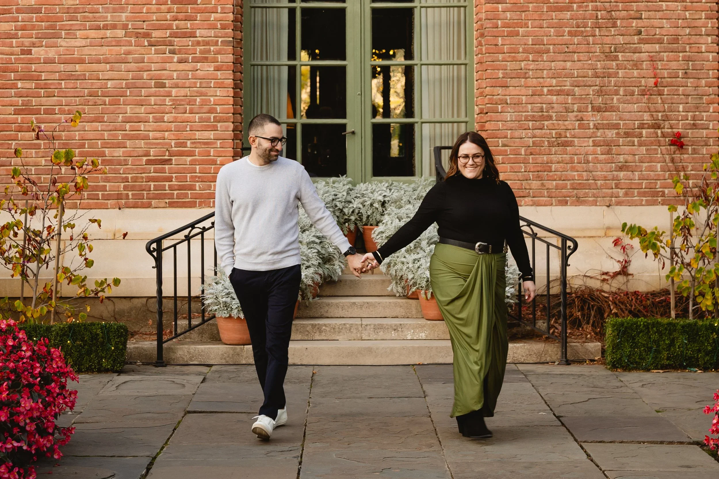 Couple walking hand in hand down the garden steps at Filoli, her olive skirt trailing behind her and pink begonias blooming in the foreground.