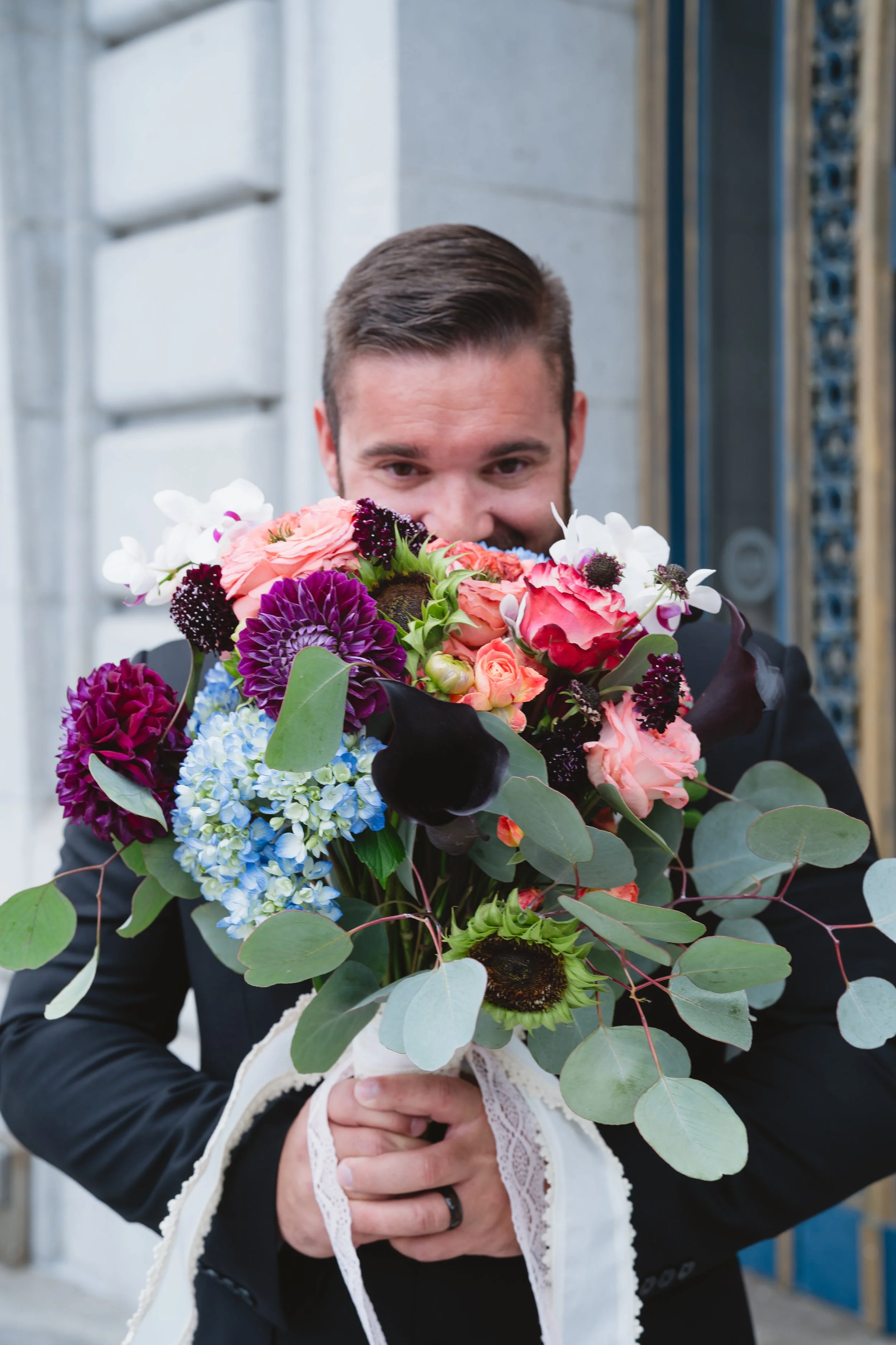 Groom peeking over a bouquet of peonies, dahlias, and hydrangea in shades of coral, purple, and blue.