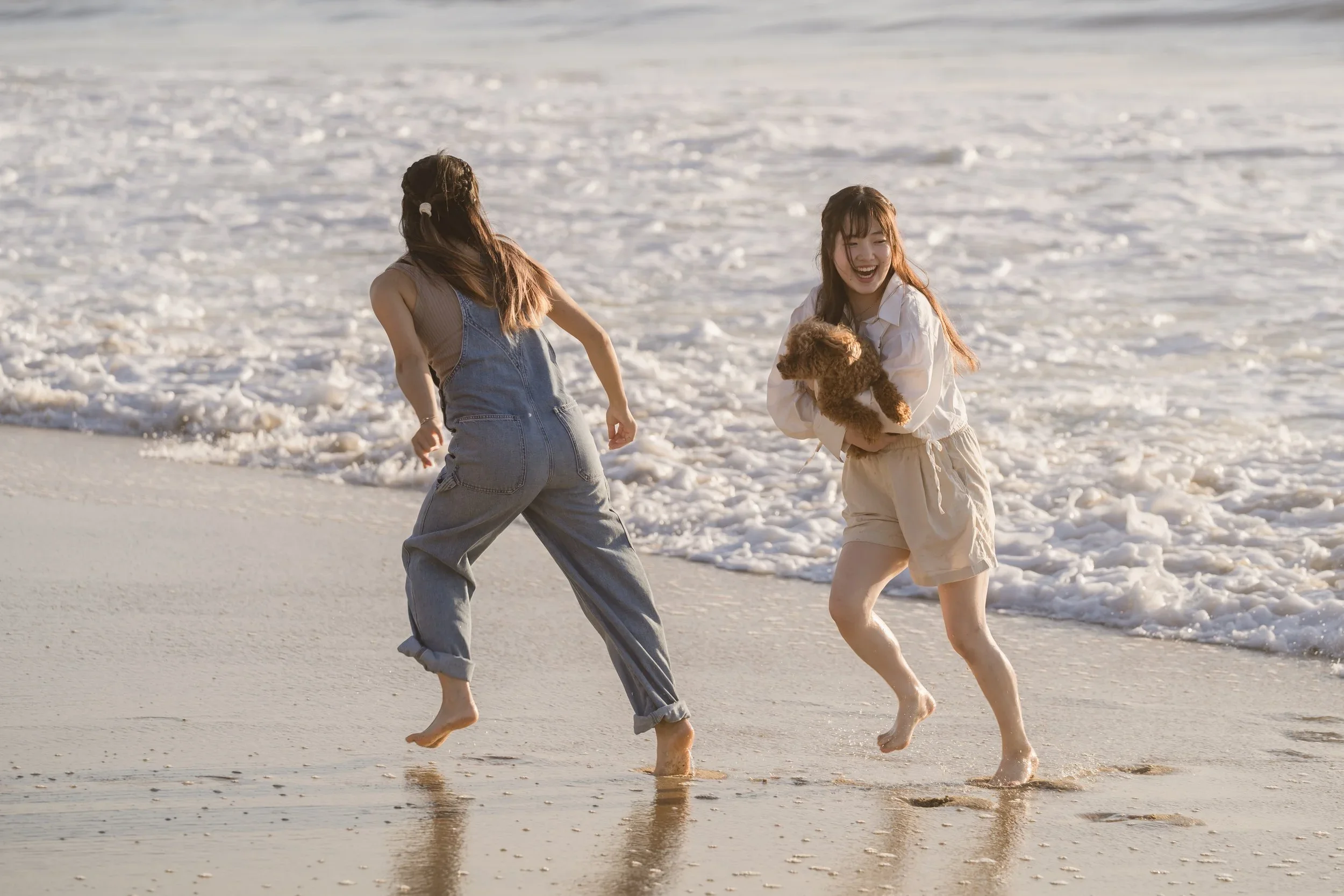 Two sisters move through shallow foam at the water's edge, one carrying the poodle mix while the other twists away from an incoming wave