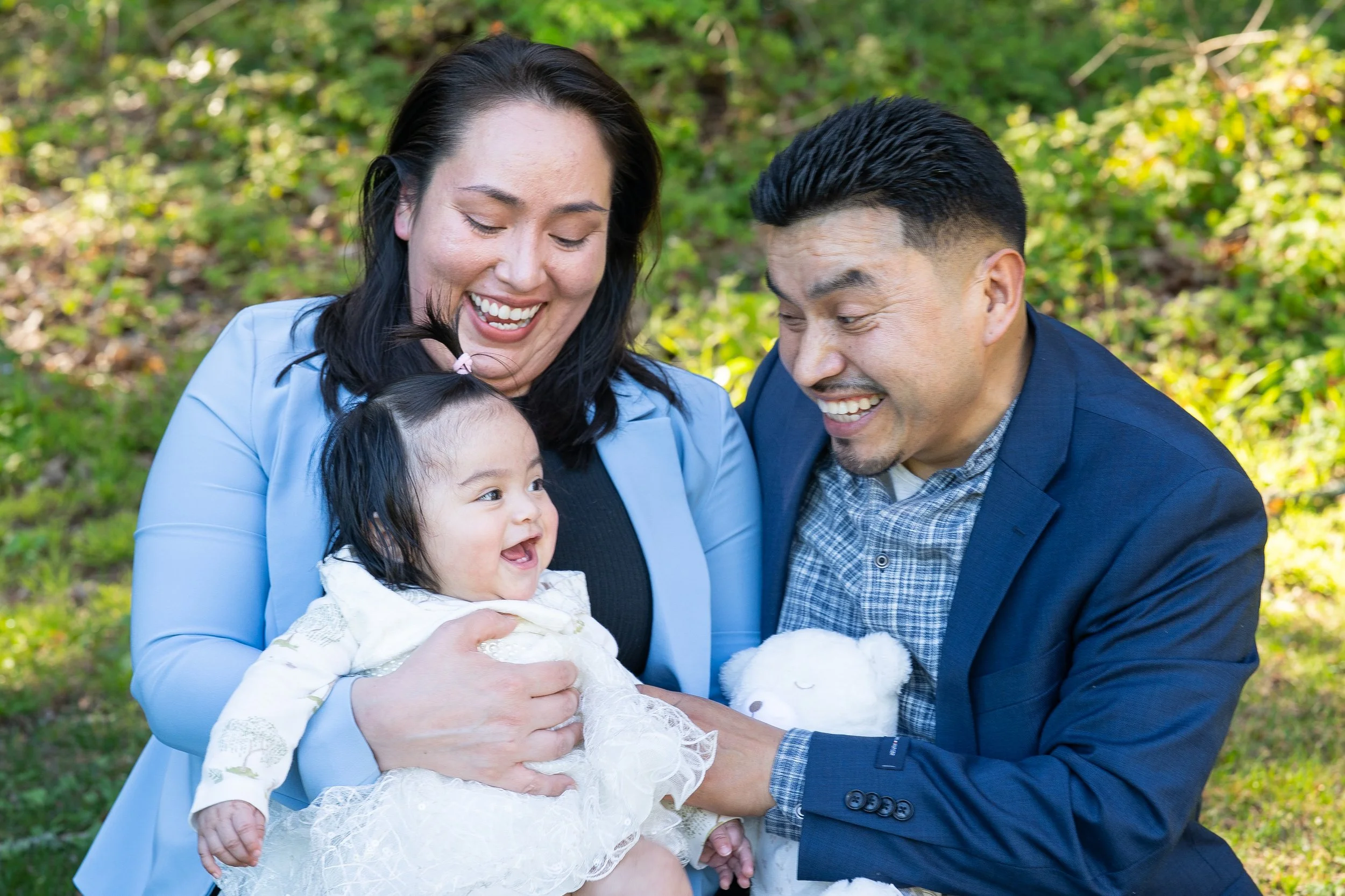 A family of three smiling outdoors in a green, wooded area. The woman has long dark hair, the man has short black hair, and they are holding a happy young girl with curly dark hair and a white lace dress.