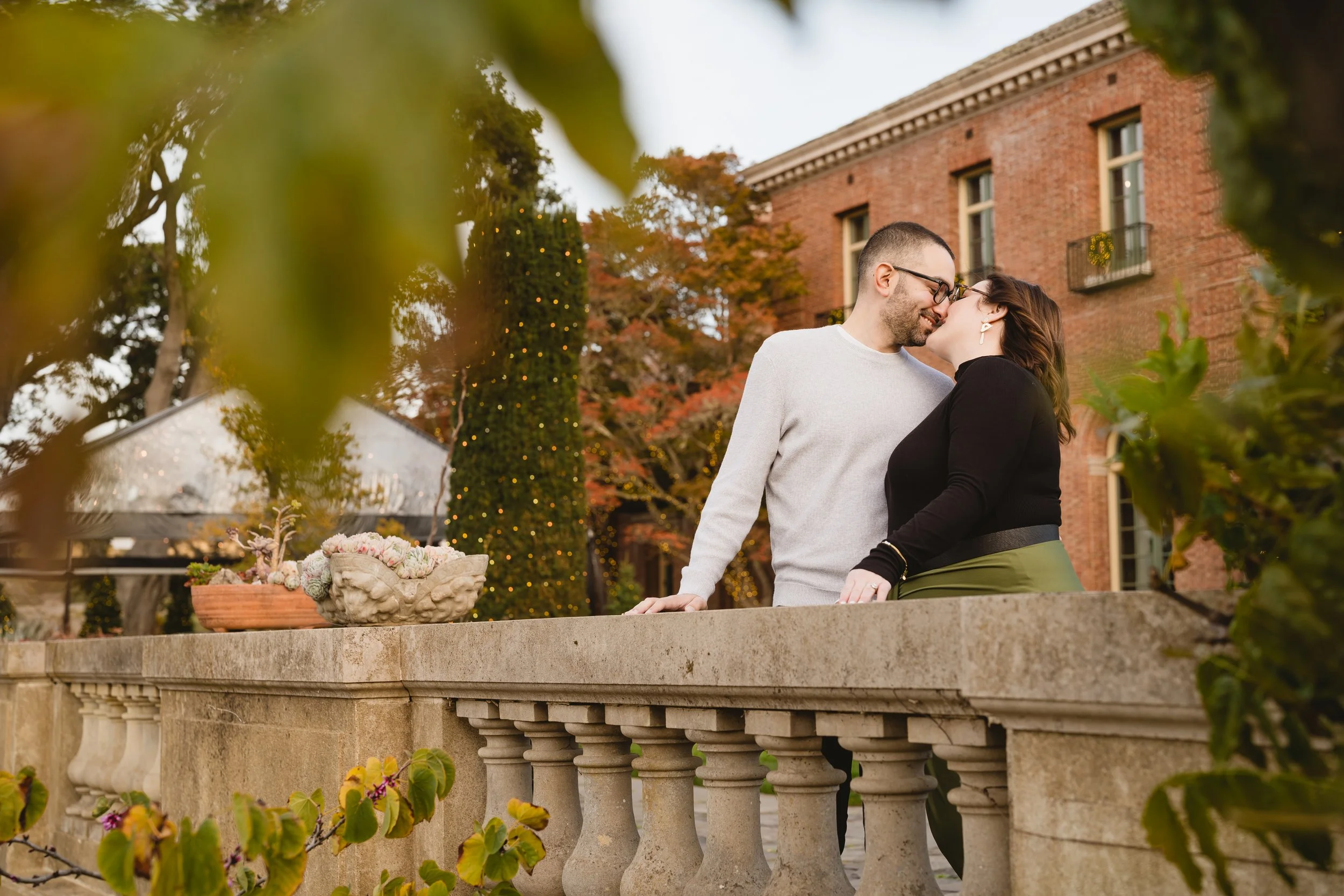 A couple sharing a kiss on an outdoor stone balcony in front of a brick building with multiple windows and a small balcony with a wreath. There are trees with fall foliage and a decorated Christmas tree in the background.