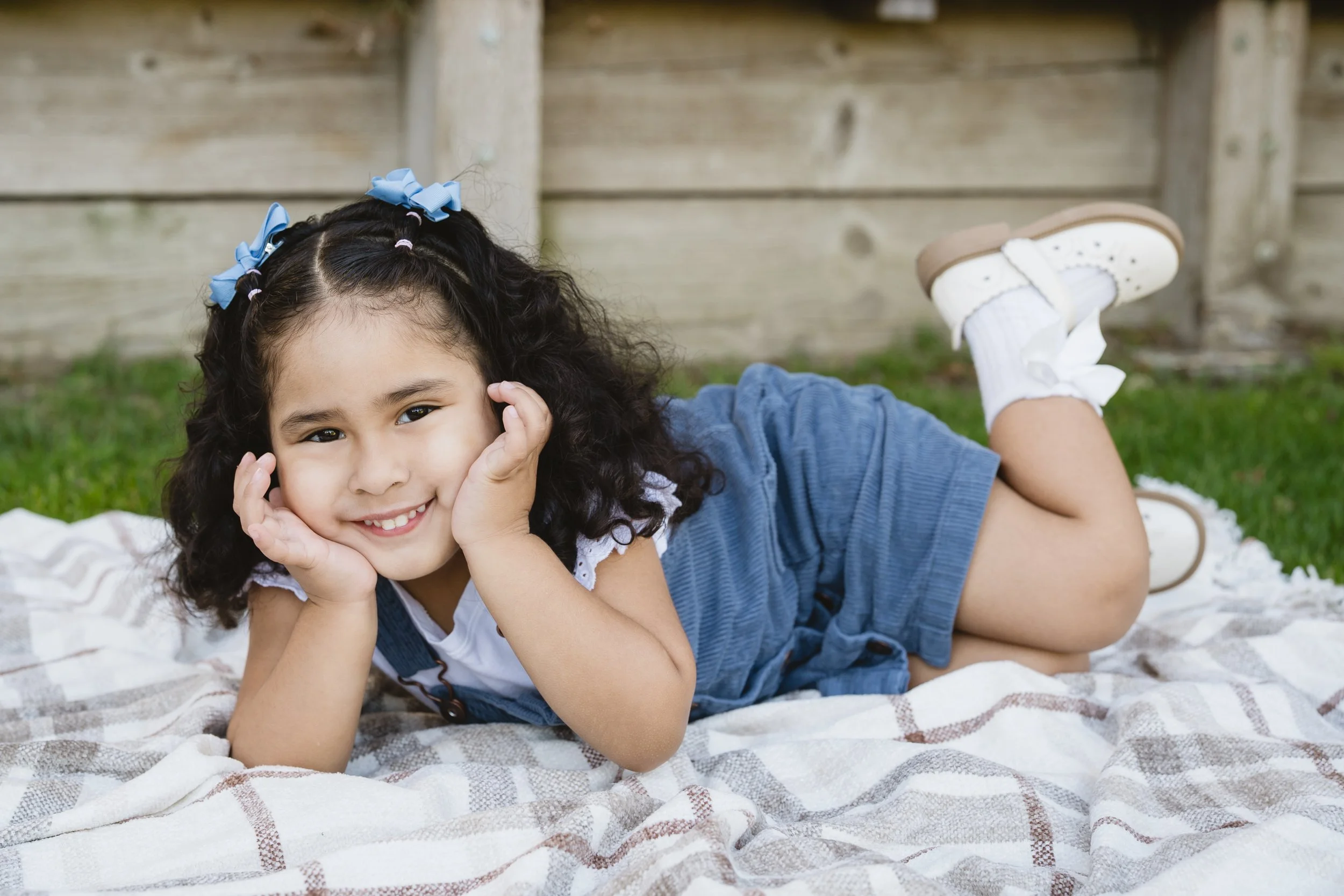A young girl with curly dark hair lying on a blanket outdoors, smiling with her hands on her cheeks, wearing a blue dress and white sneakers with socks, with a wooden fence in the background.