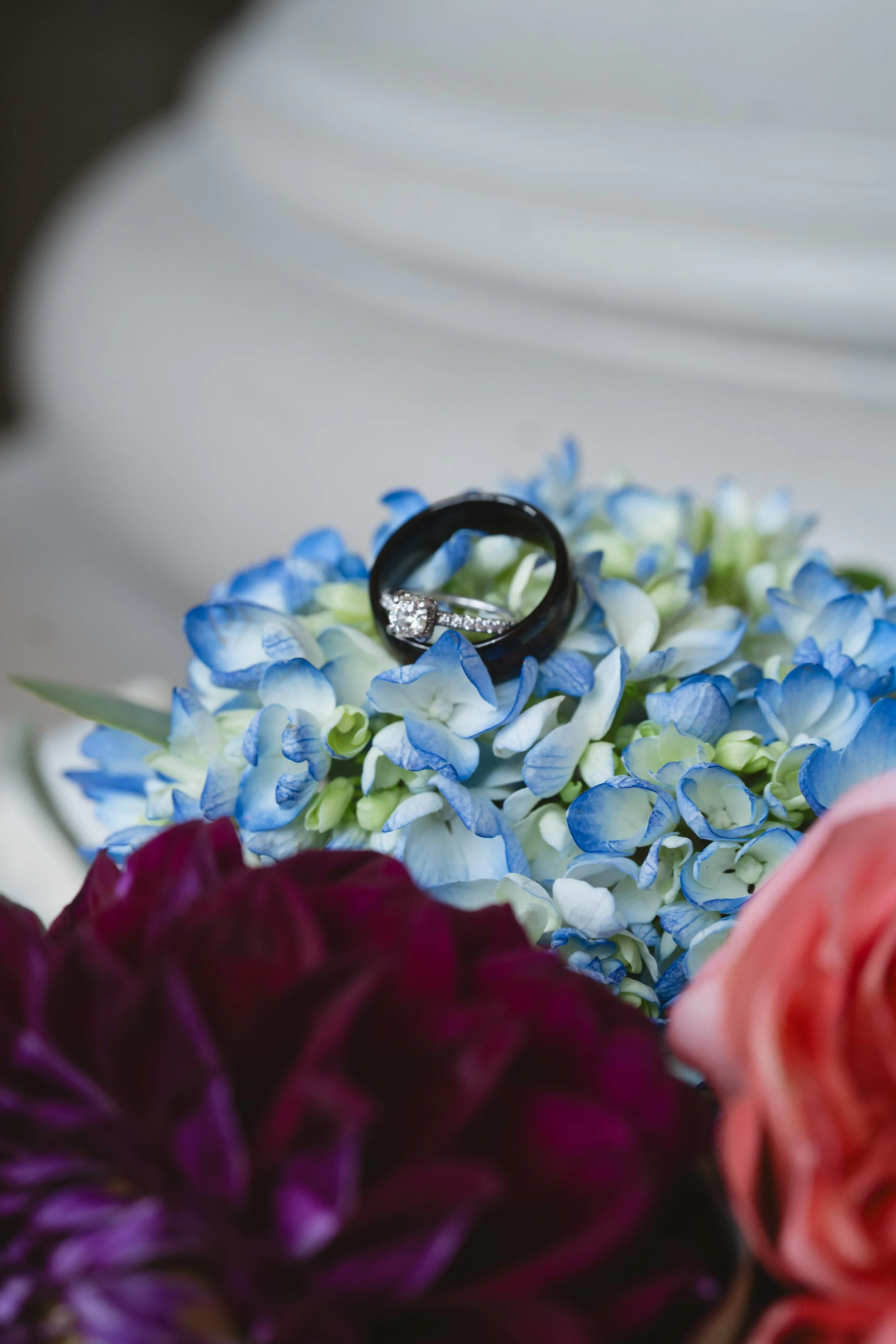 Wedding rings resting on blue hydrangea blossoms within a bouquet of coral and deep purple flowers.