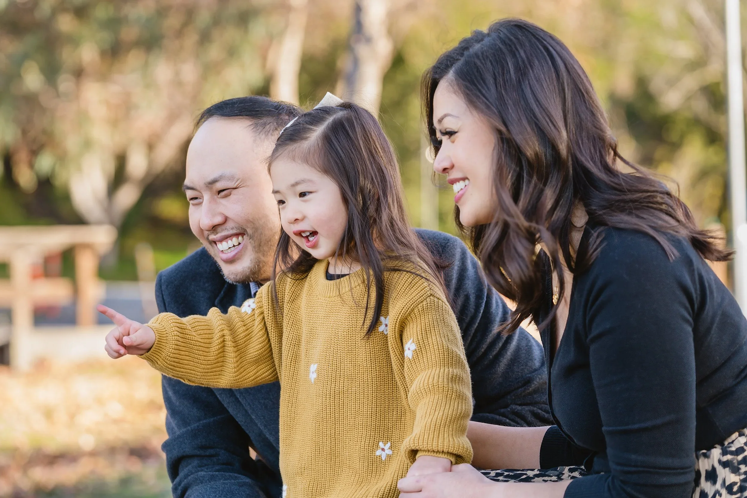In a close, candid crop, a father laughs beside his daughter as she points at something off-camera, with her mother leaning in smiling behind them.