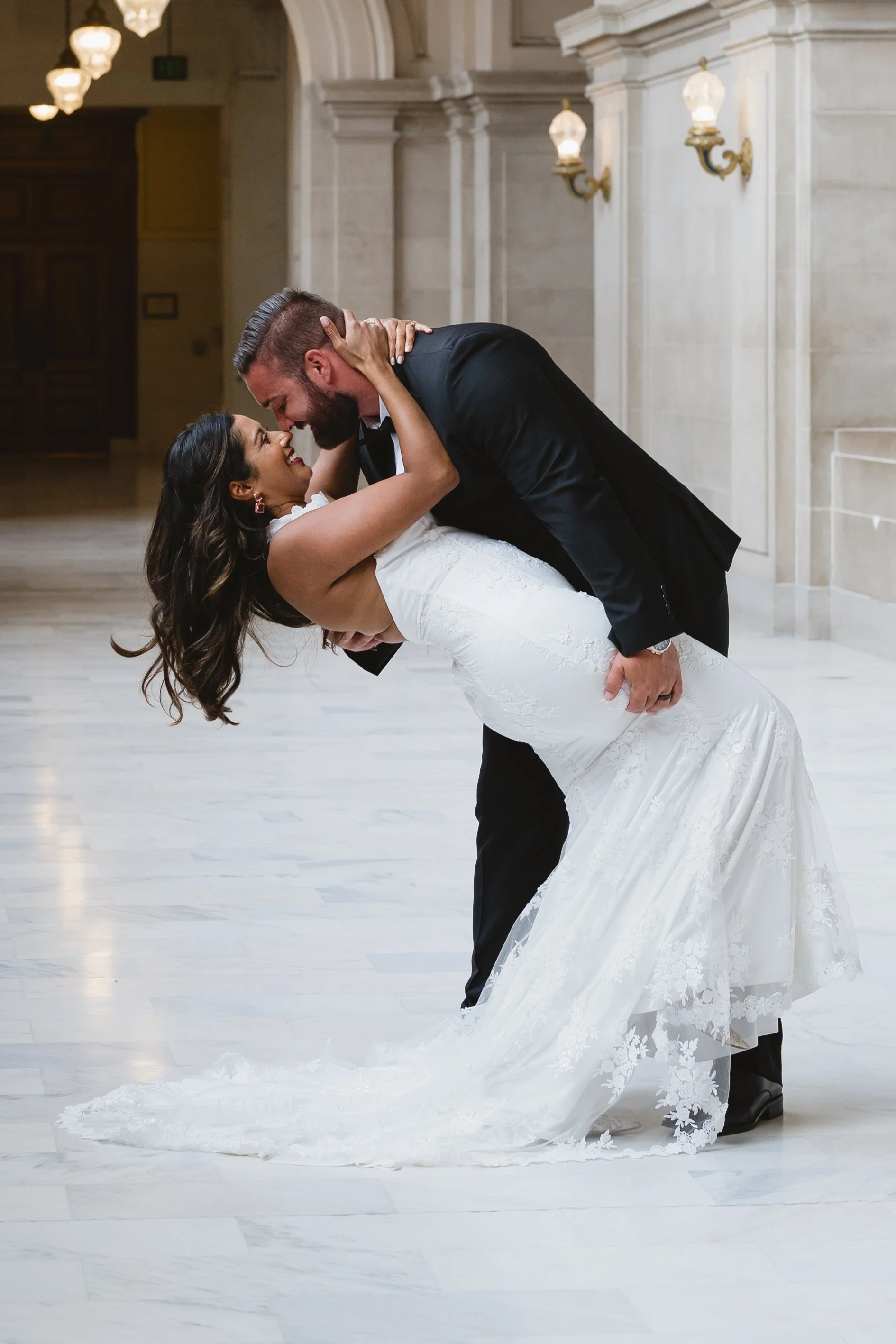 Groom dipping his bride into a kiss in a marble corridor lit by wall sconces.