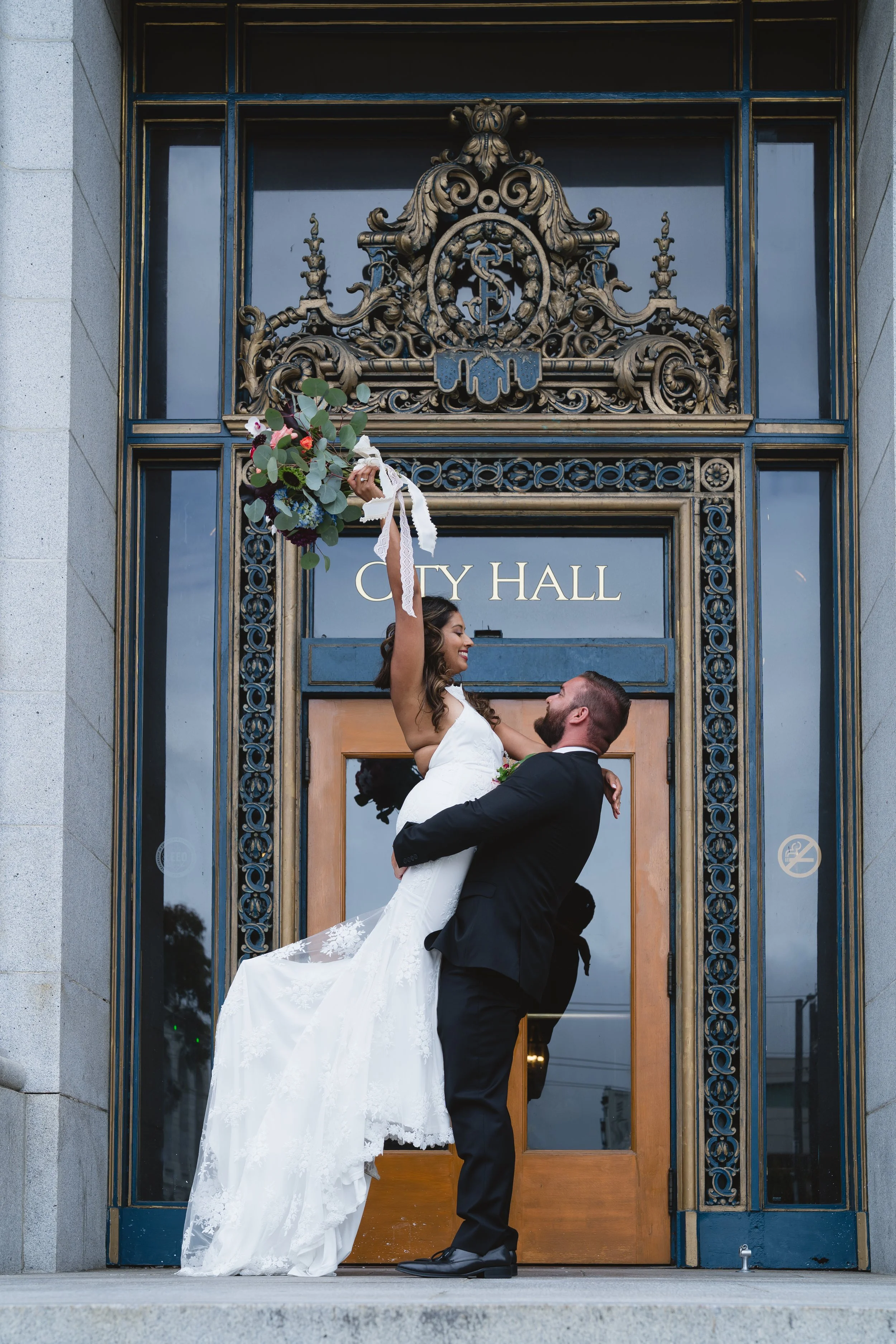 Groom lifting bride into the air beneath the ornate carved pediment above City Hall's front doors, her bouquet raised overhead.