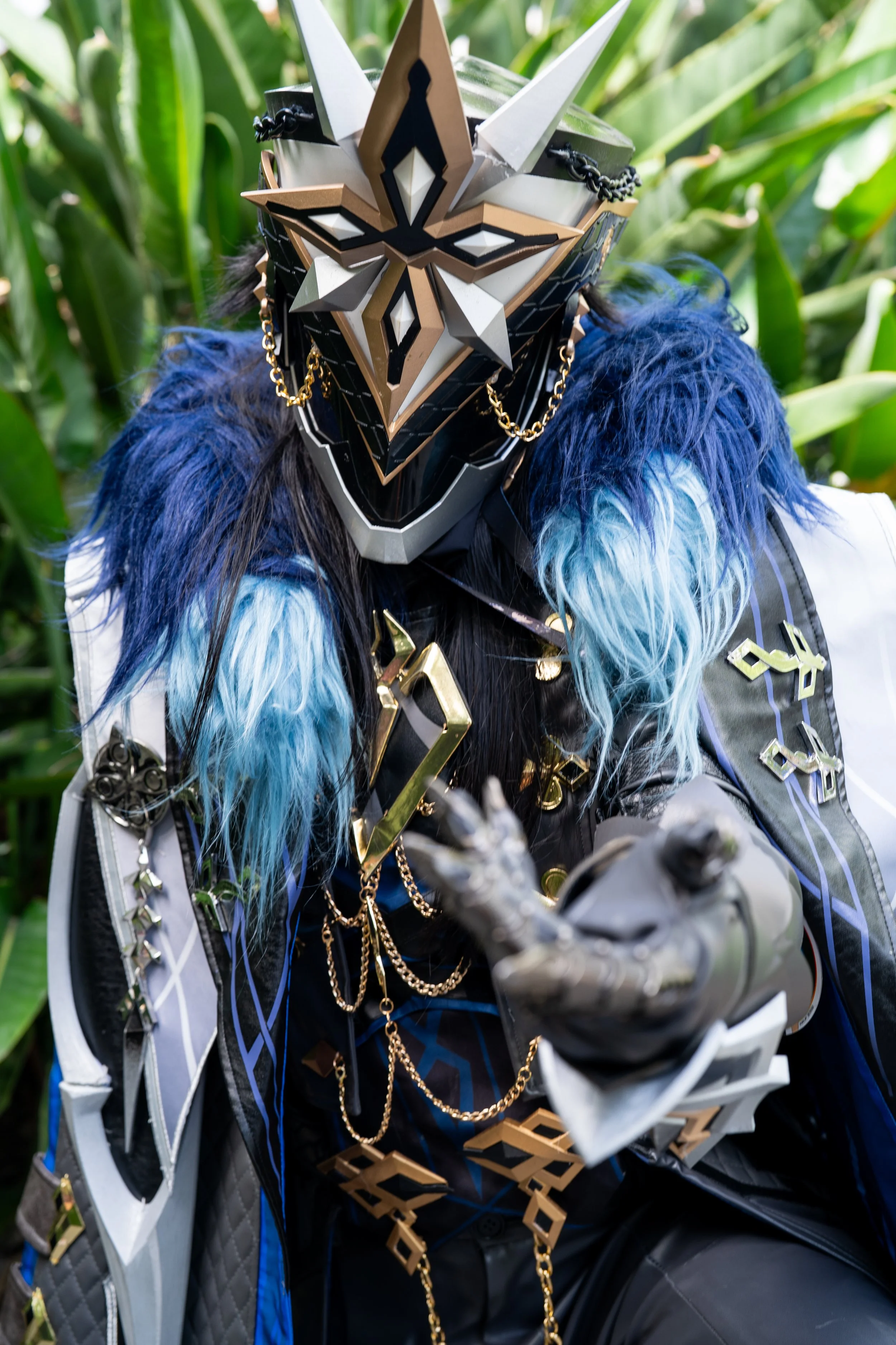 Person in an elaborate, armored costume with a dragon mask and colorful feathers, standing outdoors among green plants.