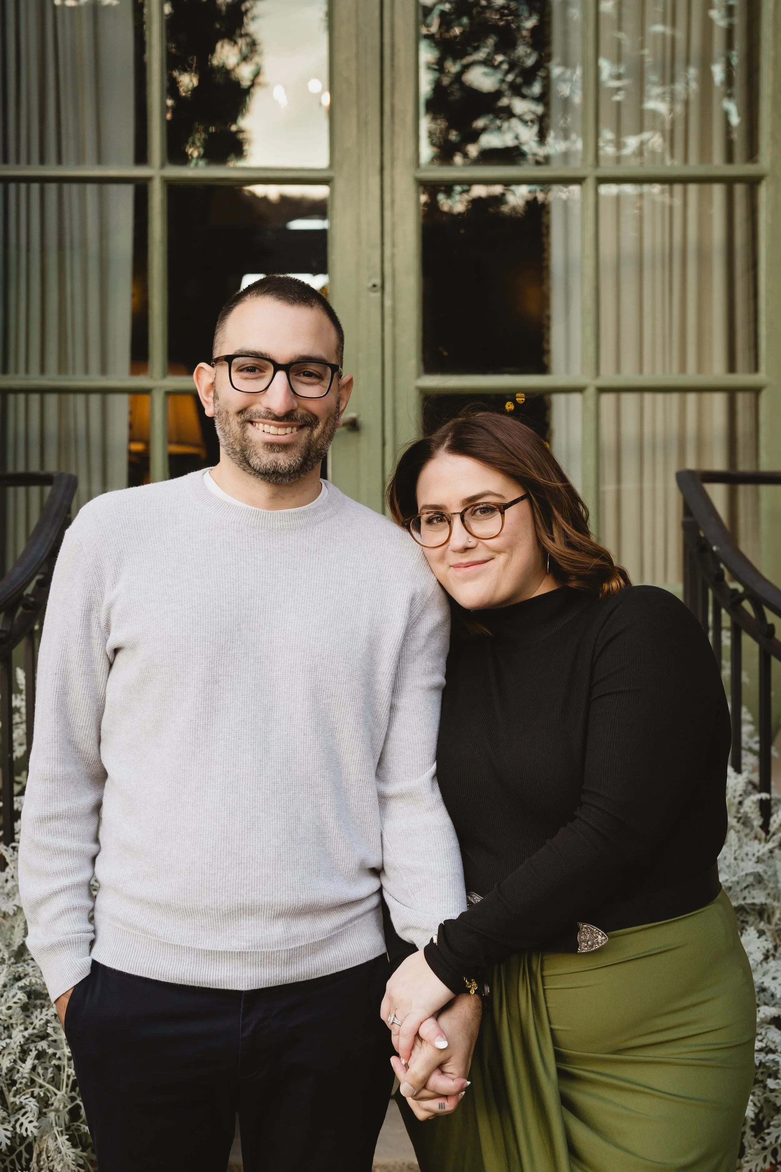 Engaged couple standing together holding hands in front of sage green garden windows, her head tipped toward his shoulder, ring visible.