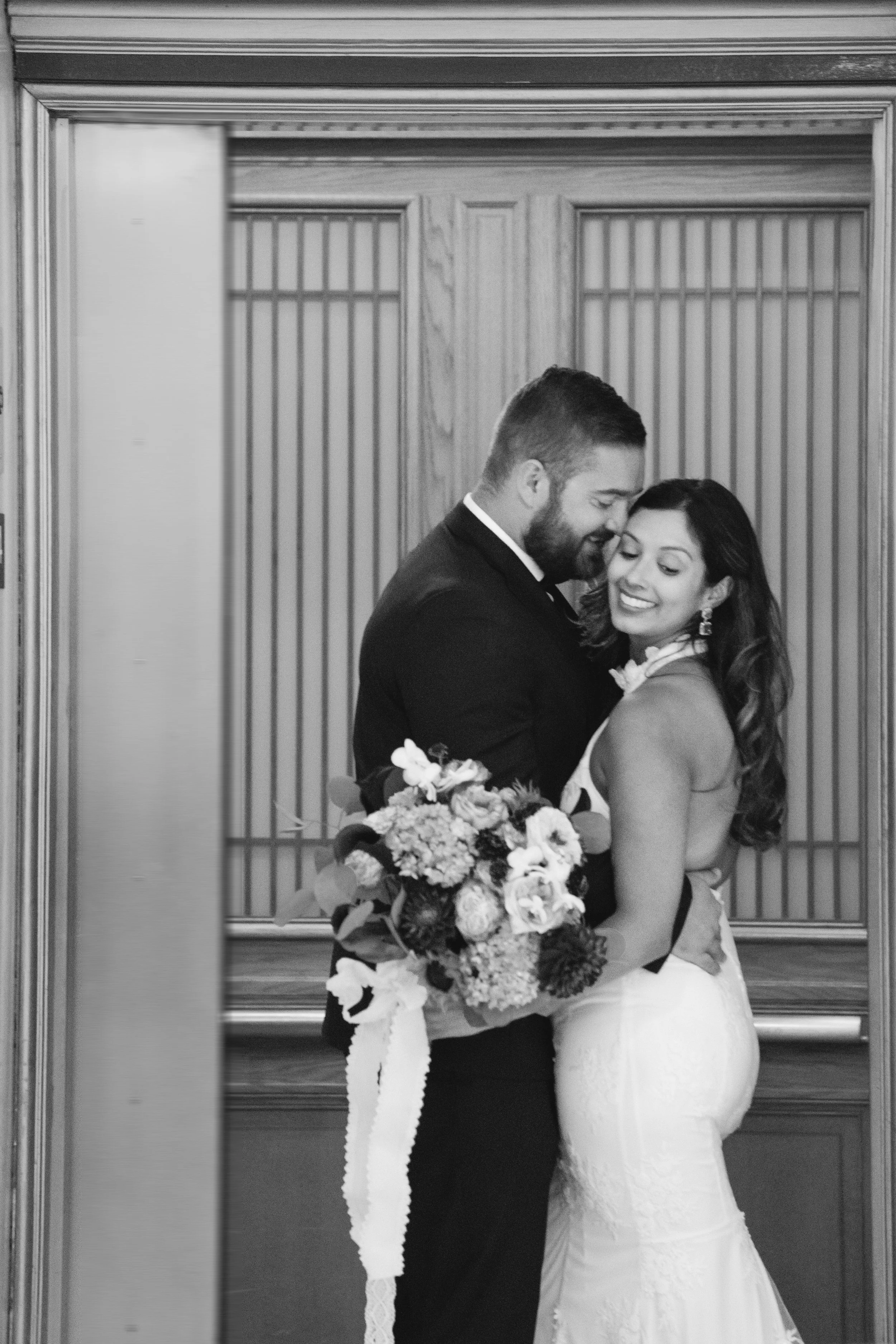 Black-and-white image of a couple embracing in front of wood-paneled elevator doors, bride glancing back over her shoulder.