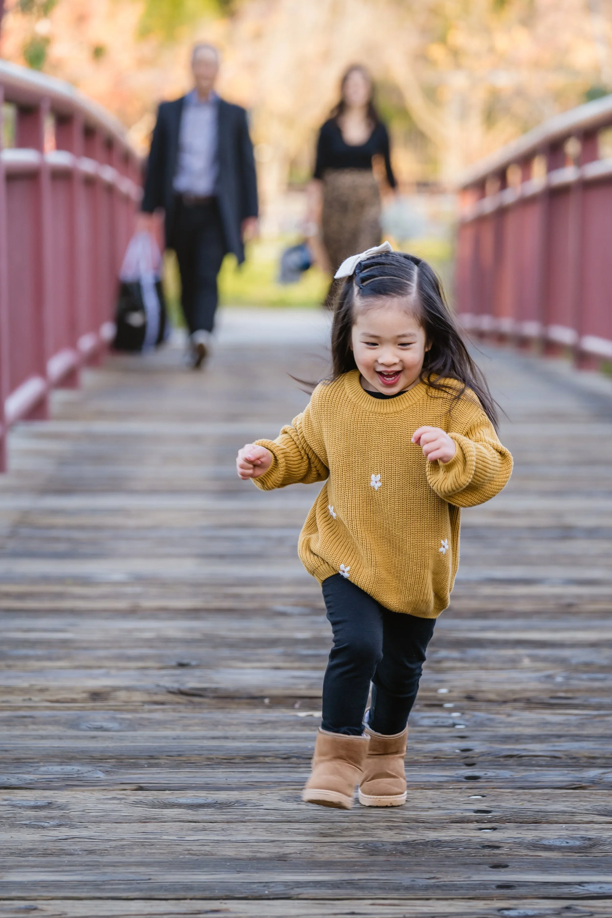 A young girl in a mustard sweater runs laughing toward the camera across a red wooden bridge, her parents walking together blurred in the background.