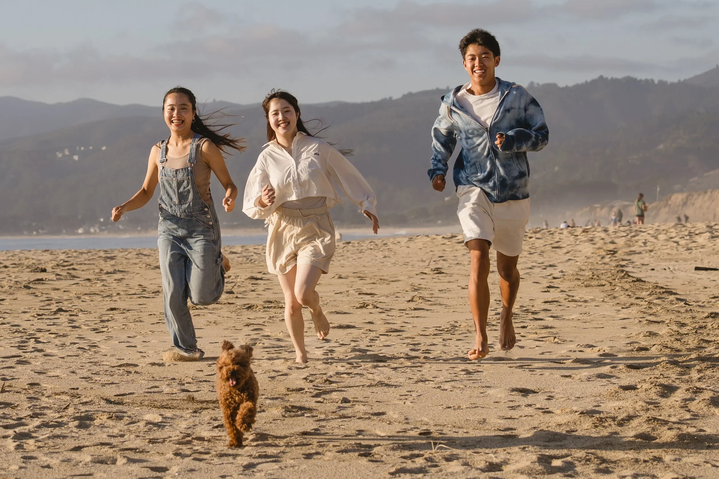Three siblings run barefoot toward the camera across wide open sand while their small brown poodle mix races ahead of them