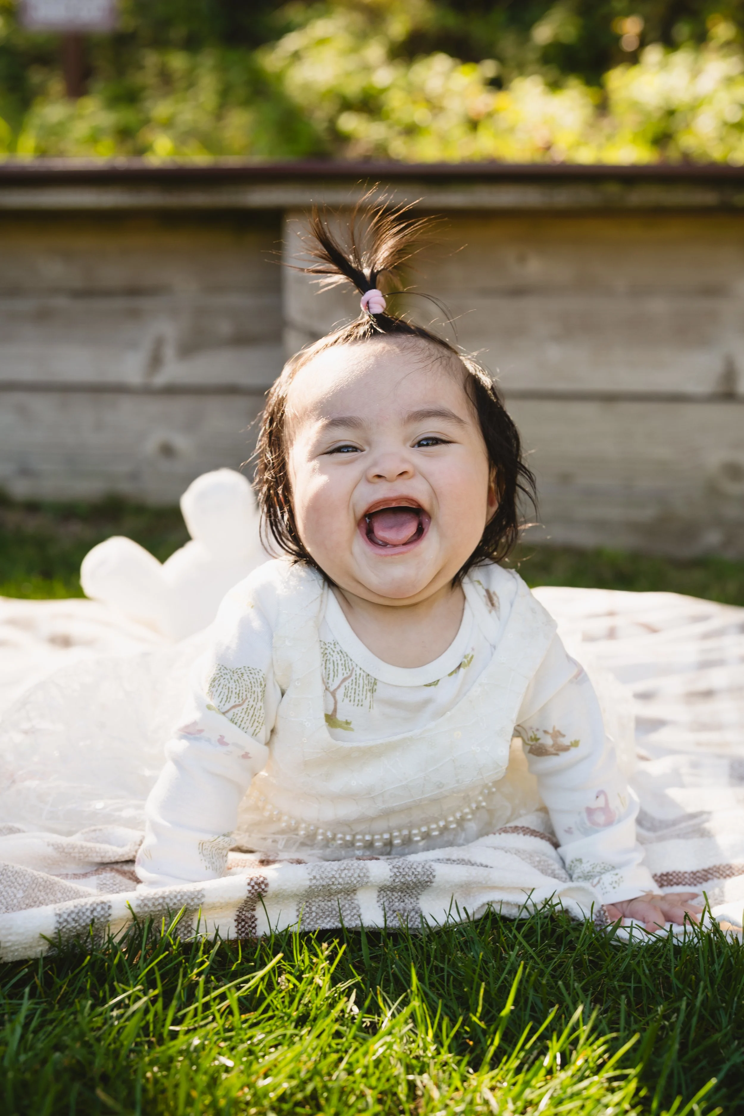 Child smiling and crawling on a blanket on grass outdoors with a wooden fence and trees in the background.