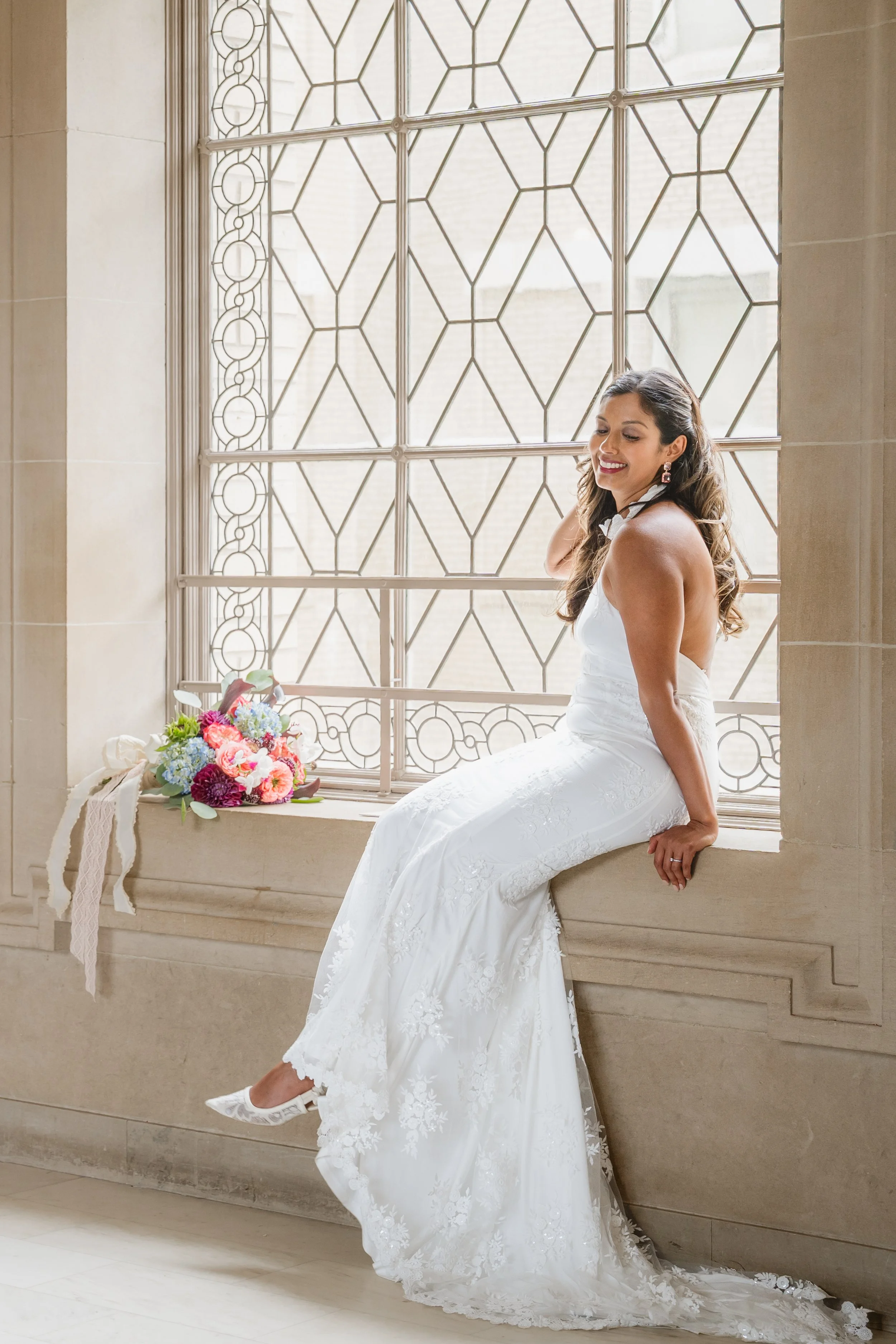Bride laughing while seated on a stone window ledge, one shoe kicked out from beneath her dress.