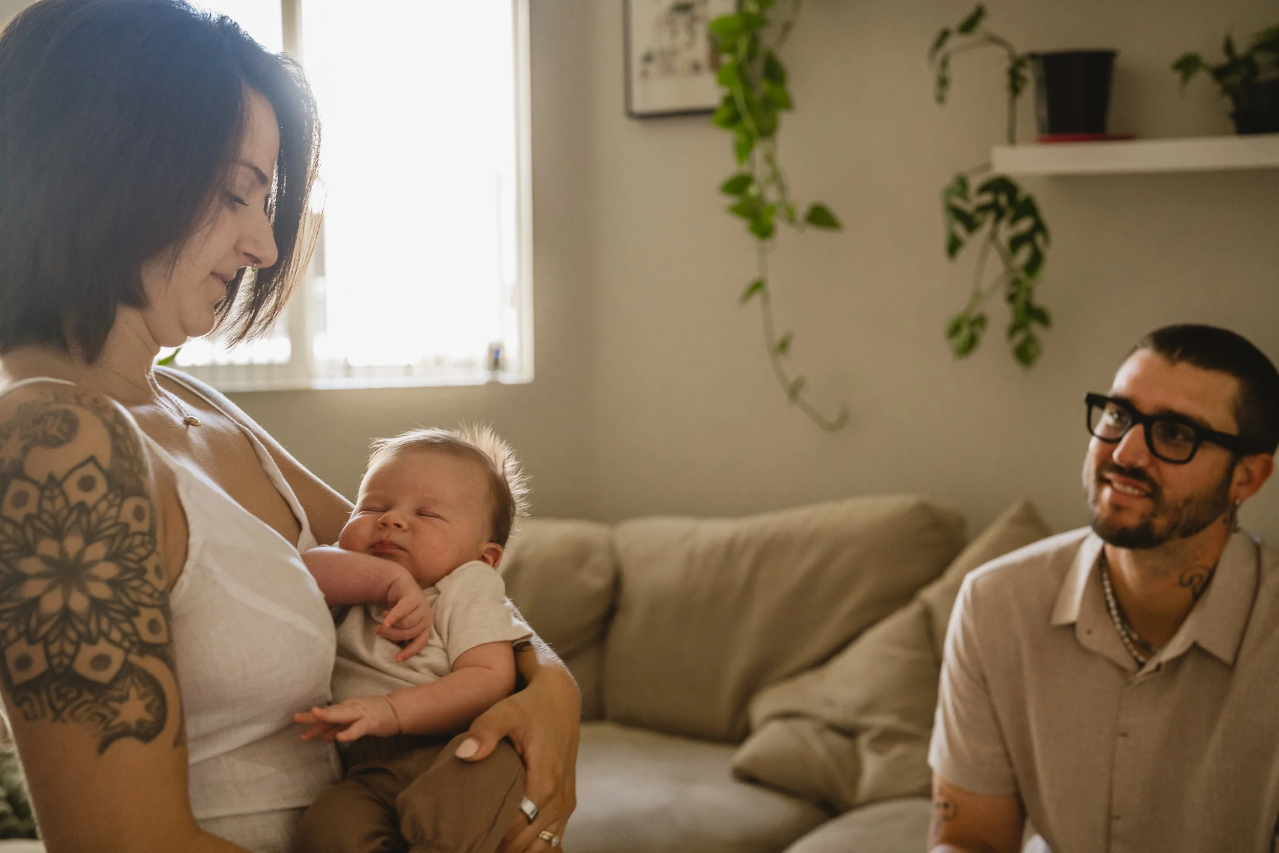 A mother stands holding her drowsy baby and looks down at them while the father, seated to the side, watches in a plant-filled living room