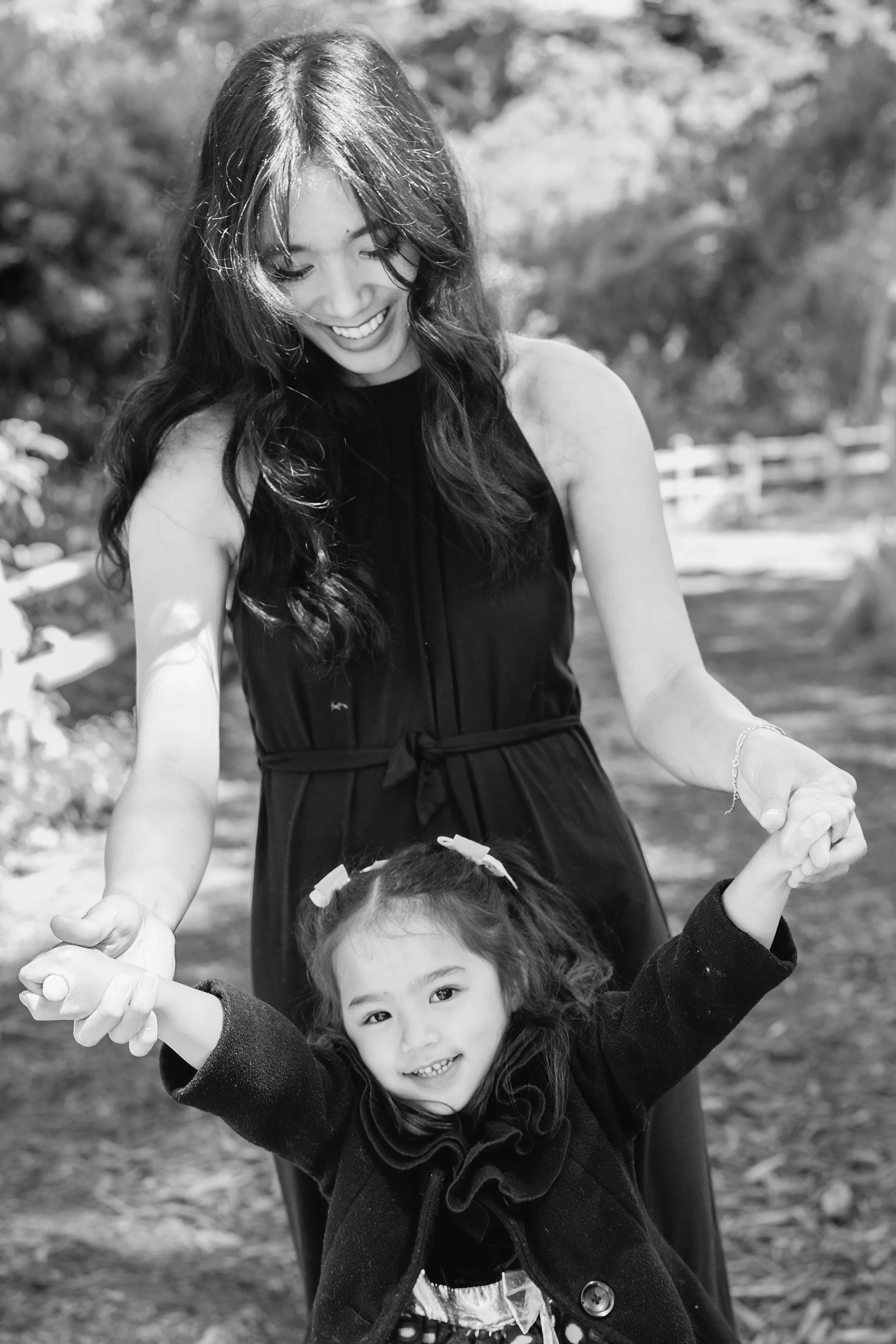 A woman and a young girl holding hands and smiling outdoors, with trees and a wooden fence in the background.