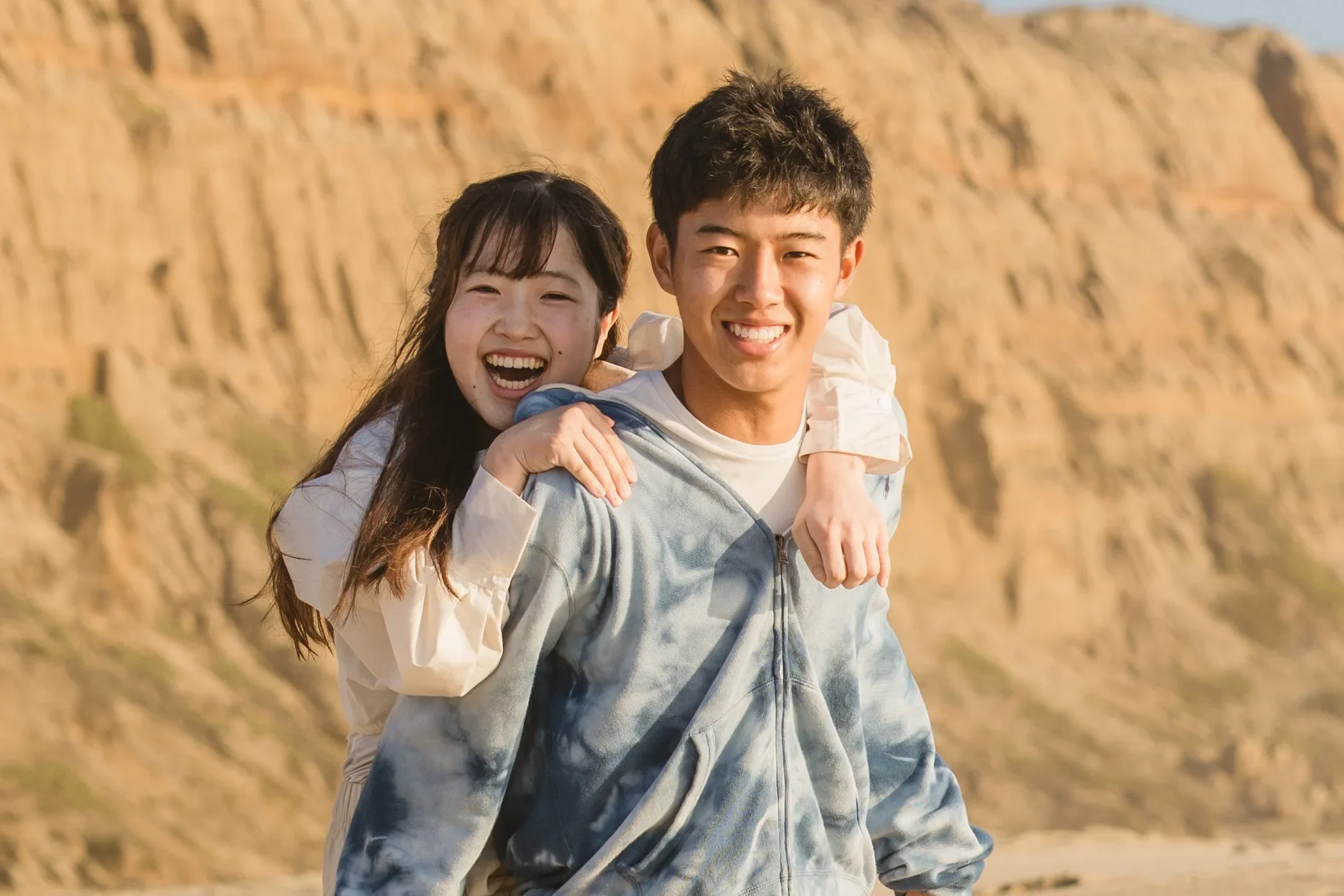 A young woman rides on her brother's back, both laughing, with the eroded sandstone cliffs of Half Moon Bay rising behind them