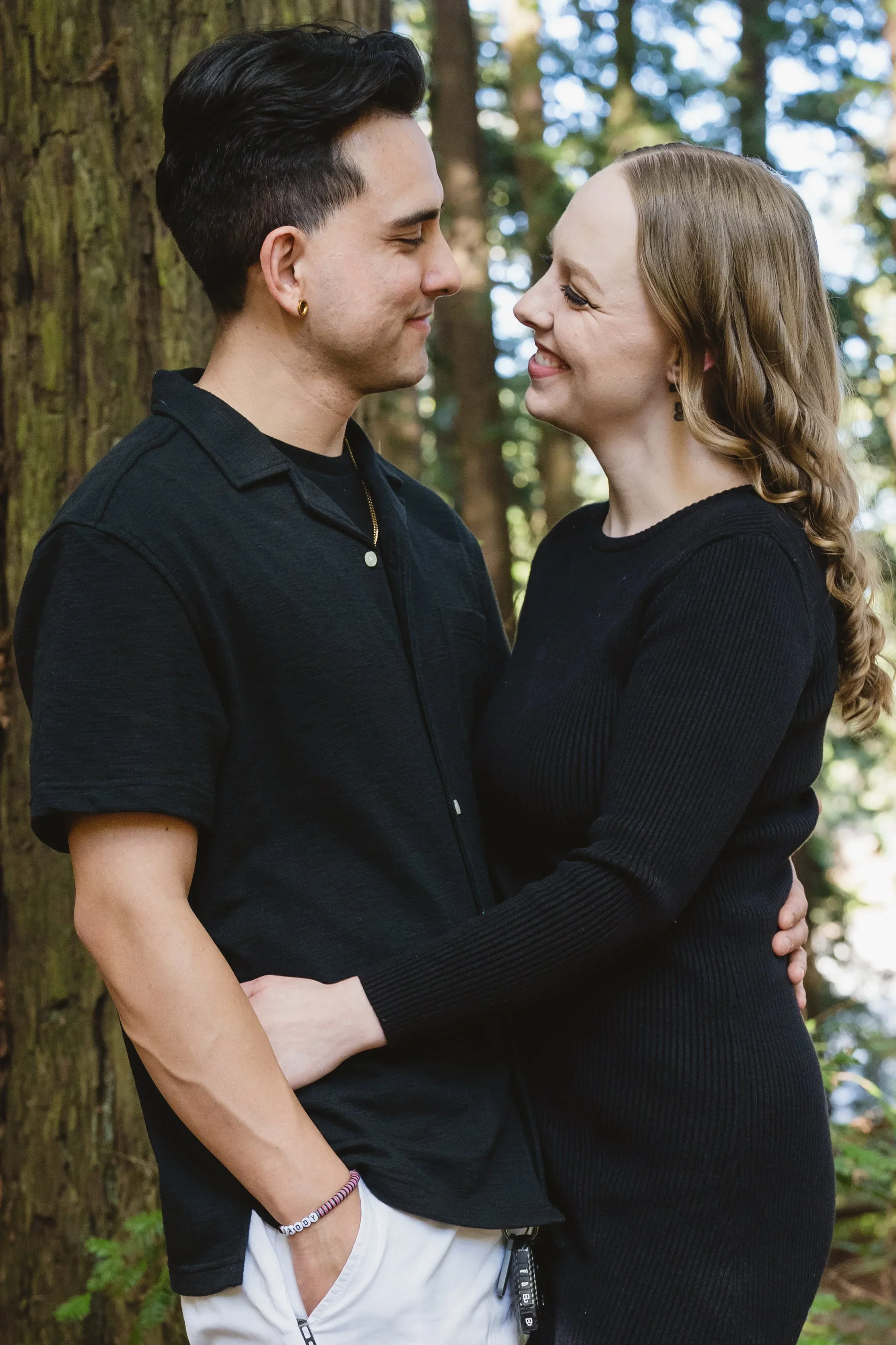 Couple standing chest to chest with their arms wrapped around each other in front of a mossy redwood trunk, both in black and looking at one another