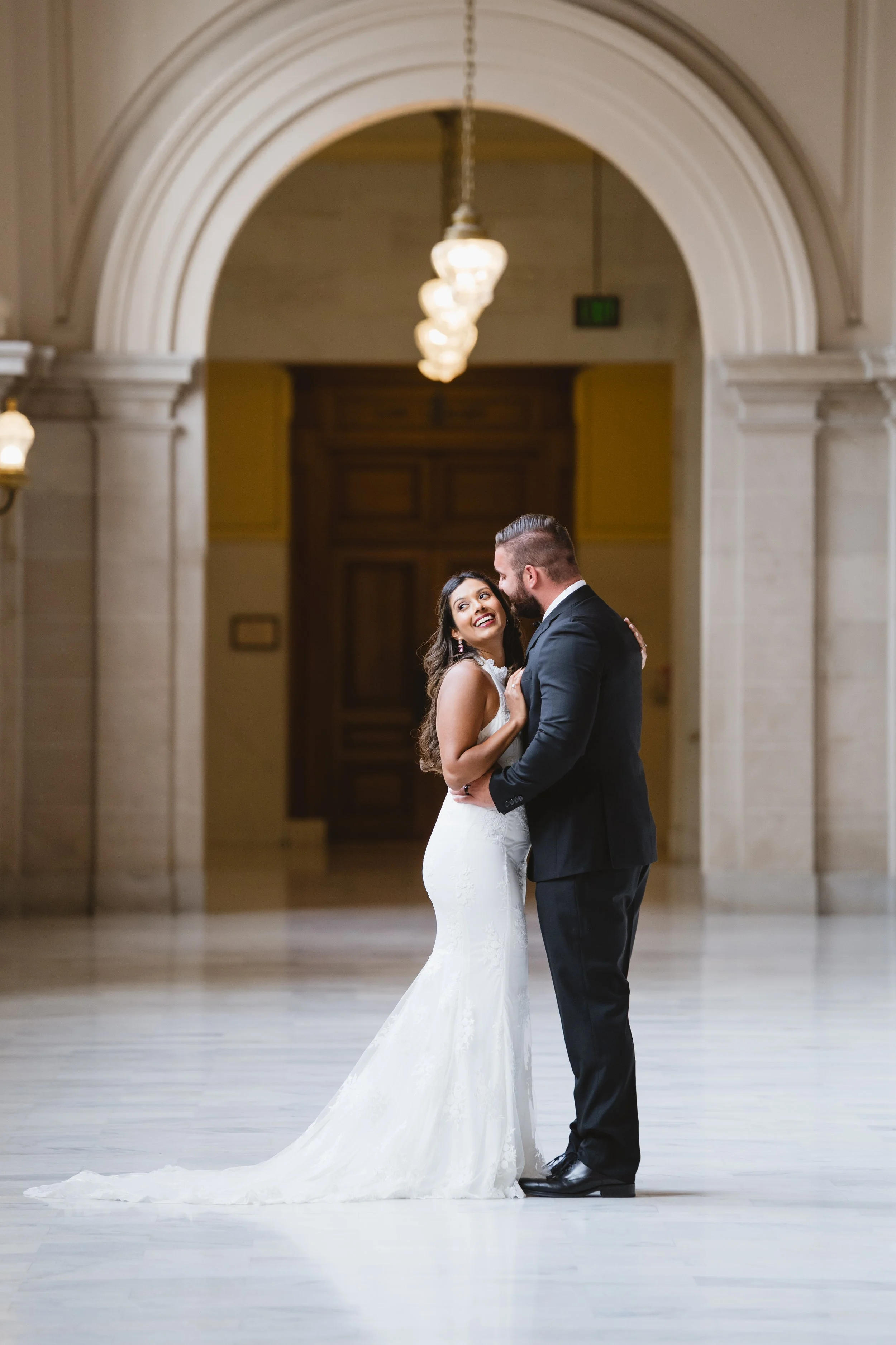 Bride glancing back over her shoulder while embracing her groom beneath a corridor archway.