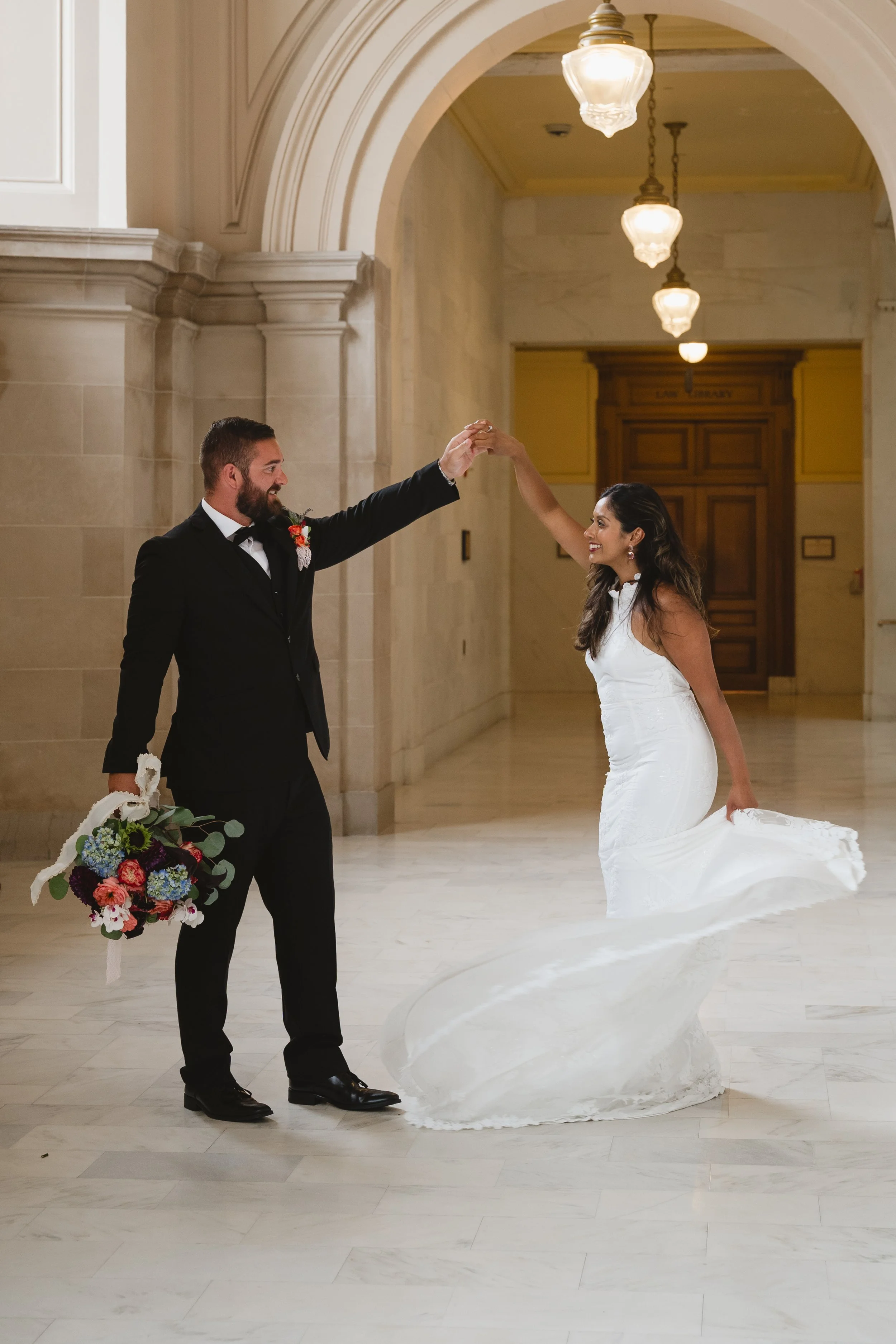 Groom twirling his bride under hanging lantern lights in a City Hall corridor, her dress swirling around her.