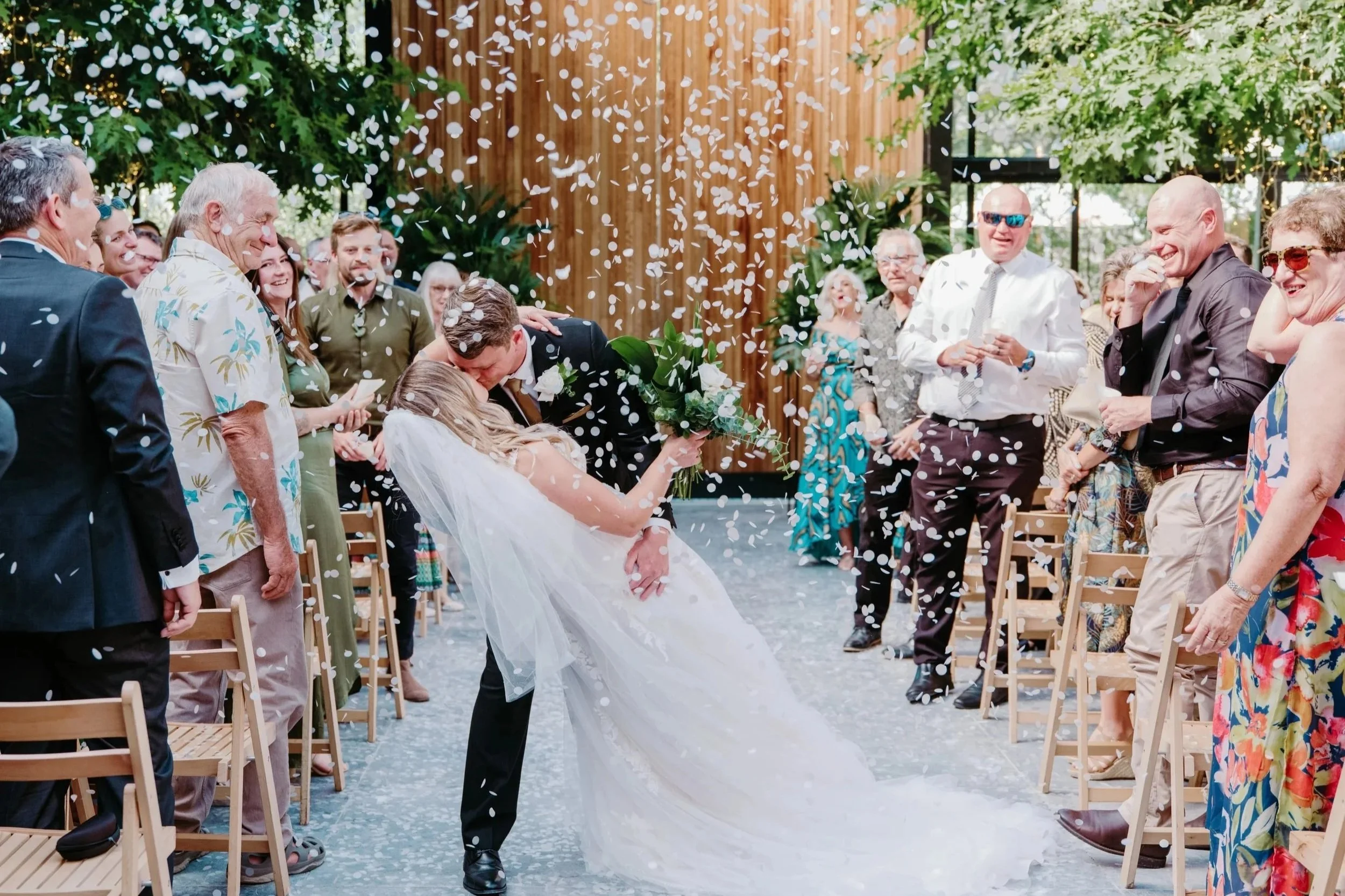A bride and groom share a romantic kiss during their outdoor ceremony as white confetti falls around them, surrounded by smiling guests seated on wooden chairs.