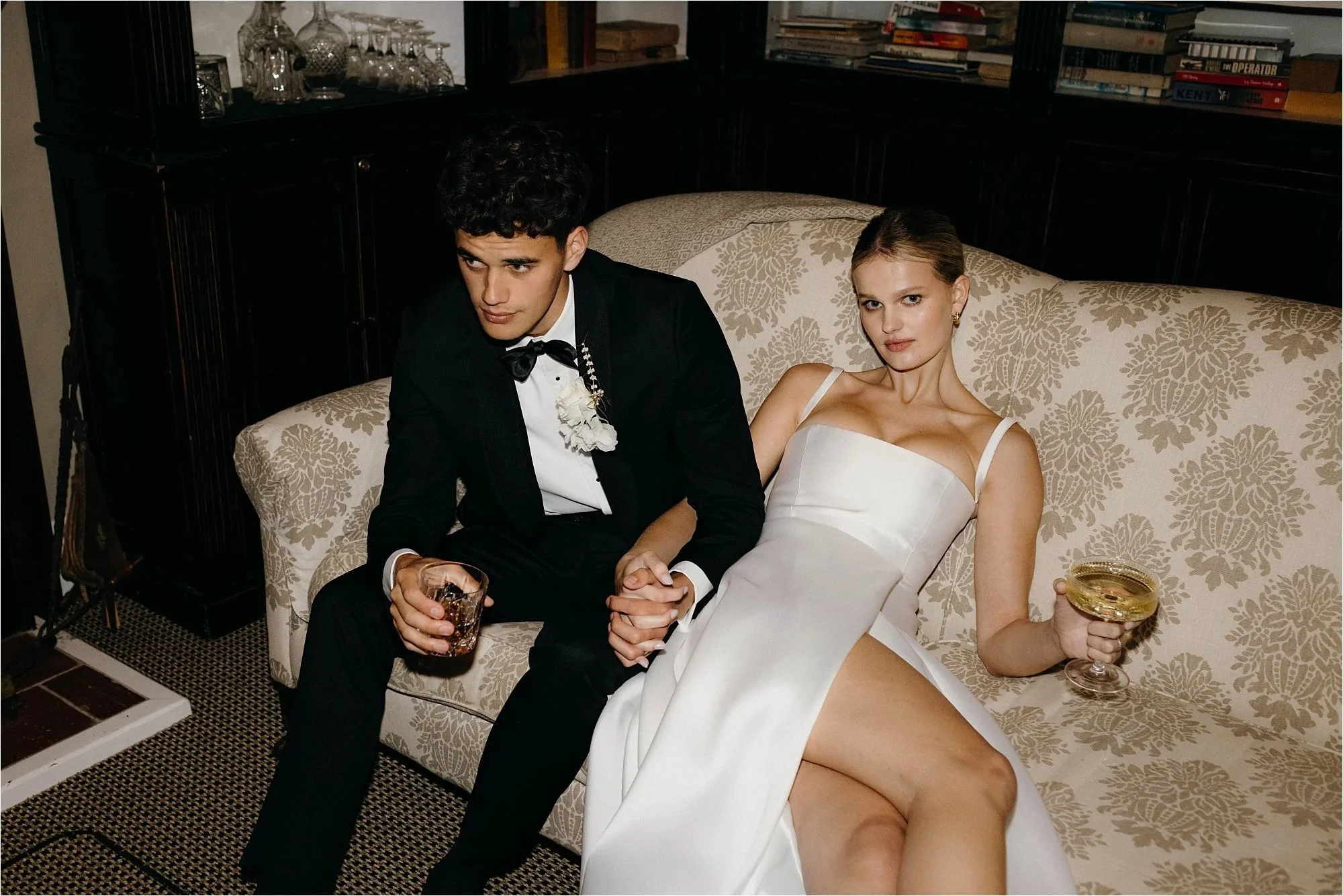 Elegant editorial wedding photo of a couple lounging on a patterned sofa with drinks, captured in a moody indoor setting.