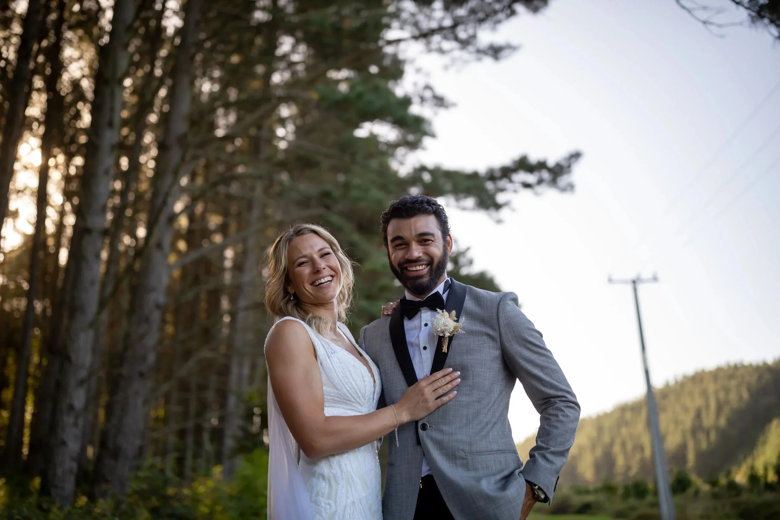 Joyful outdoor wedding portrait of a couple standing in front of sunlit trees, photographed in a candid, documentary style.