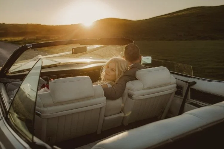 Golden-hour wedding portrait of a bride and groom in a vintage convertible, bathed in warm sunset light on a rural road.