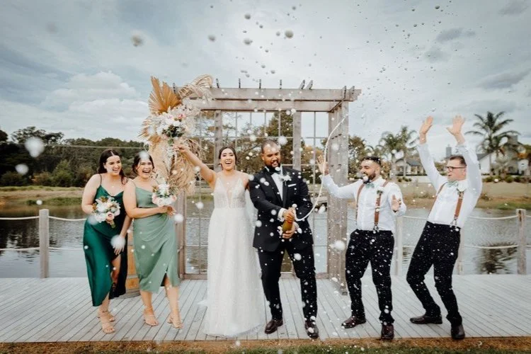 Fun outdoor wedding portrait of the bridal party cheering as the groom opens champagne beside a lakeside ceremony backdrop.