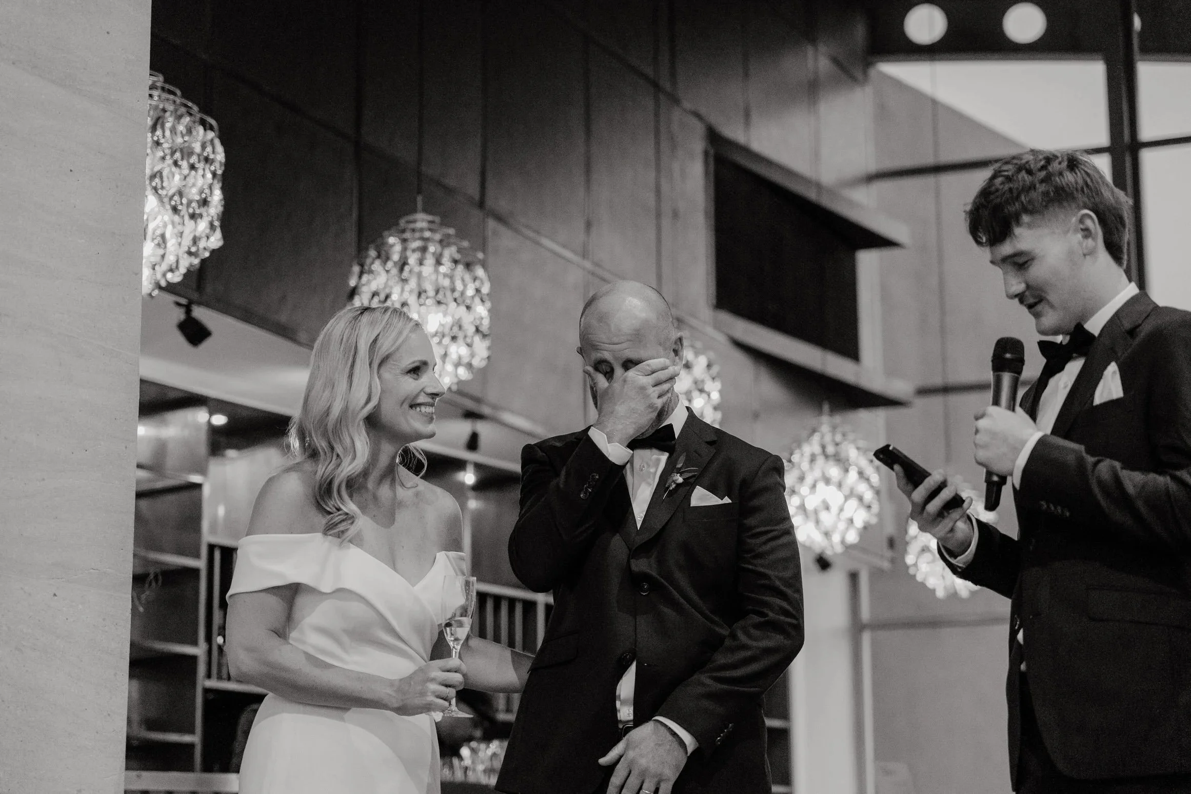 Emotive black-and-white wedding photo capturing a groom’s reaction during speeches, with the bride and a speaker beside him in a modern reception setting.