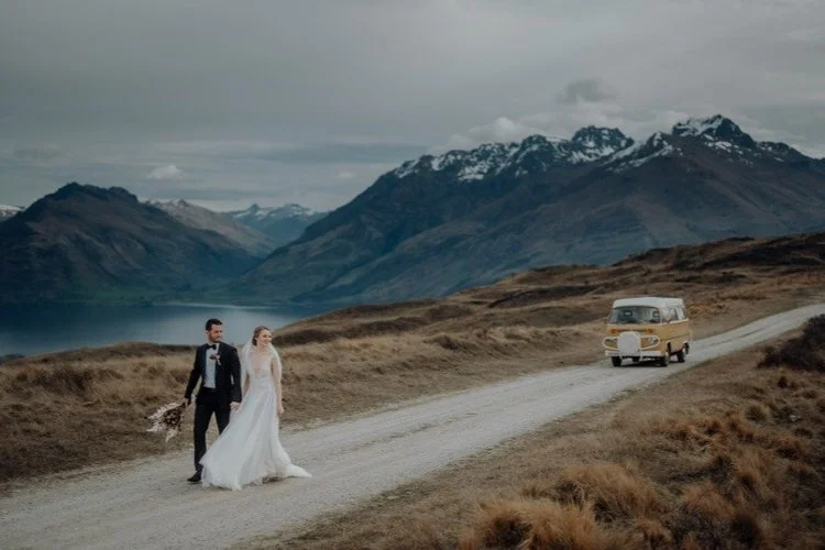 Adventure-style mountain wedding portrait of a bride and groom walking along a rural road with snow-capped peaks, lakes, and a vintage van in the distance.