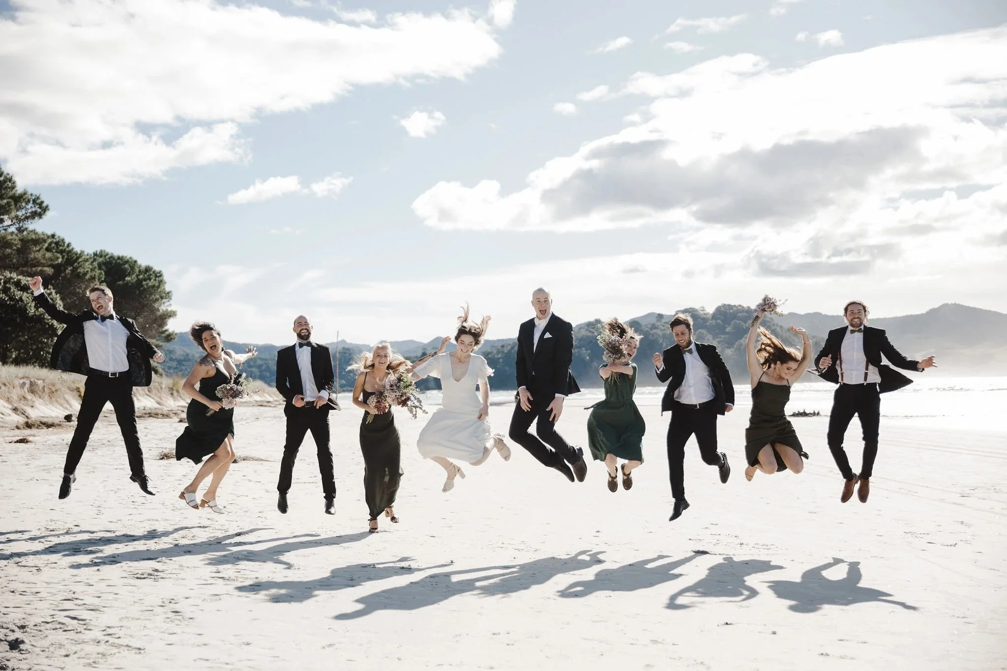 Vibrant coastal wedding image of the bridal party jumping on the sand, photographed in bright natural light with ocean and hills in the background.