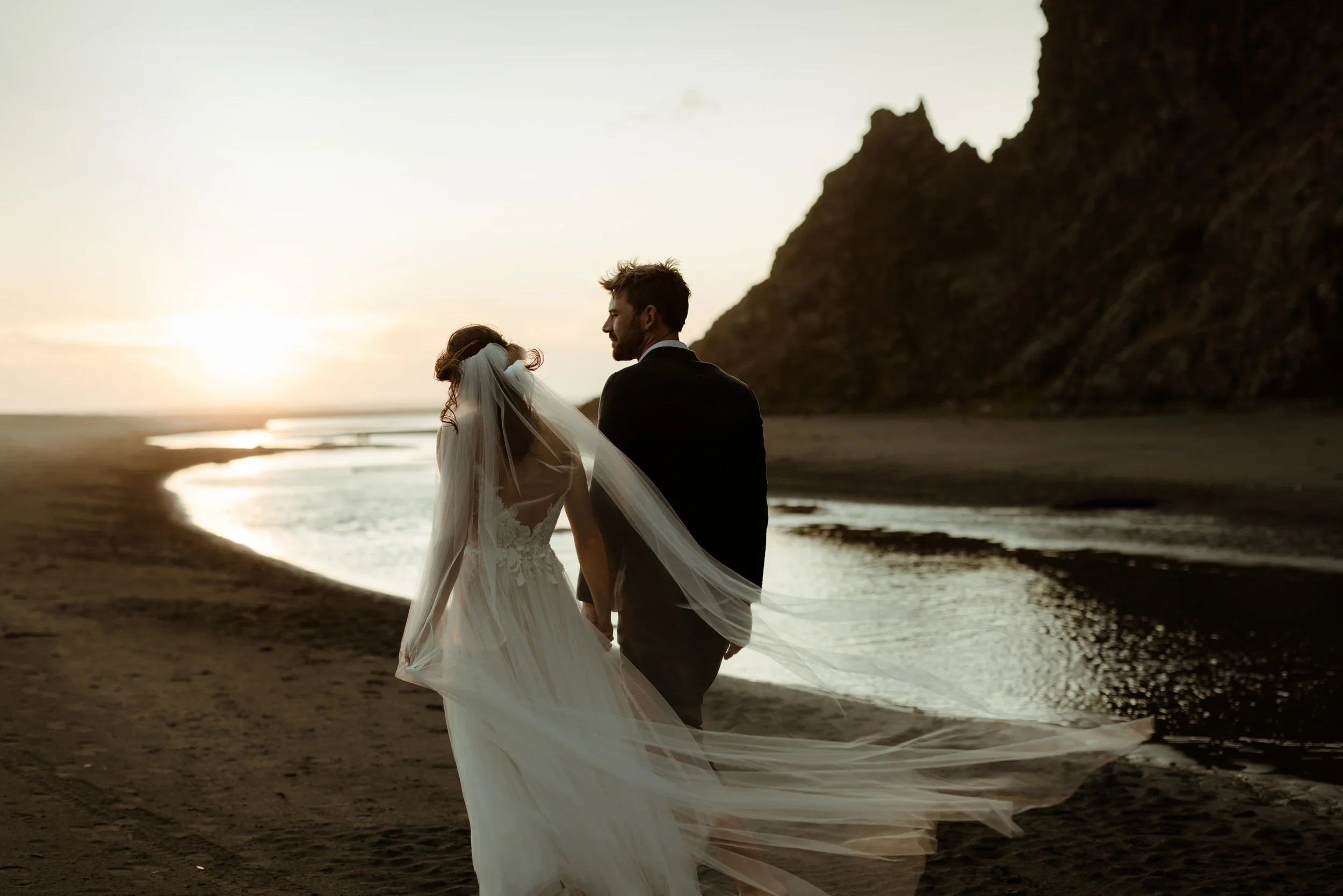 Dramatic sunset wedding portrait of a couple on a black-sand beach, captured with soft coastal light and a long, wind-blown veil.