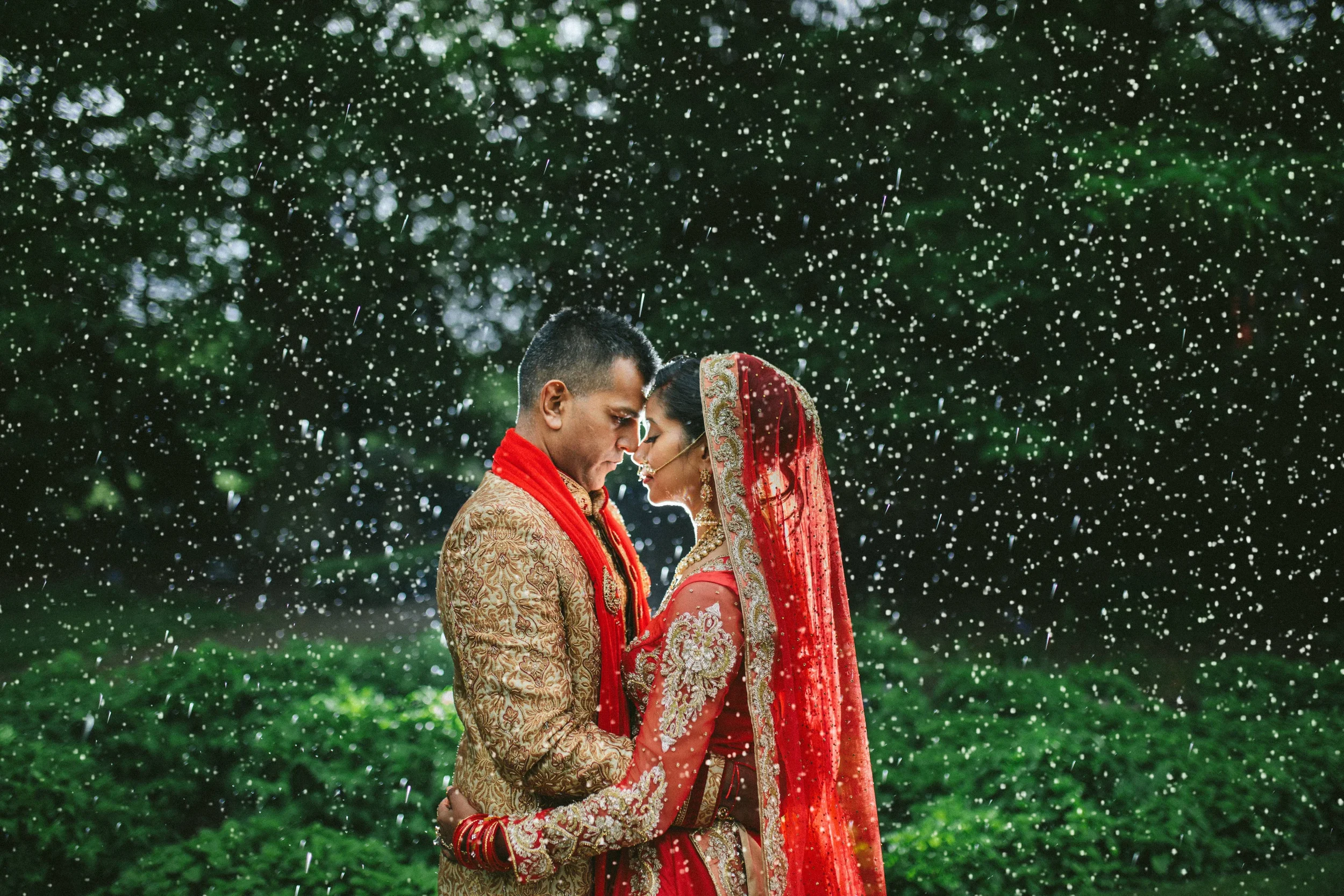 Romantic rain-soaked wedding portrait of a couple in traditional South Asian attire embracing outdoors, surrounded by falling droplets.