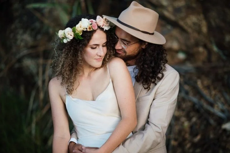 Romantic outdoor wedding portrait of a couple embracing, with the bride wearing a floral crown and the groom in a hat.