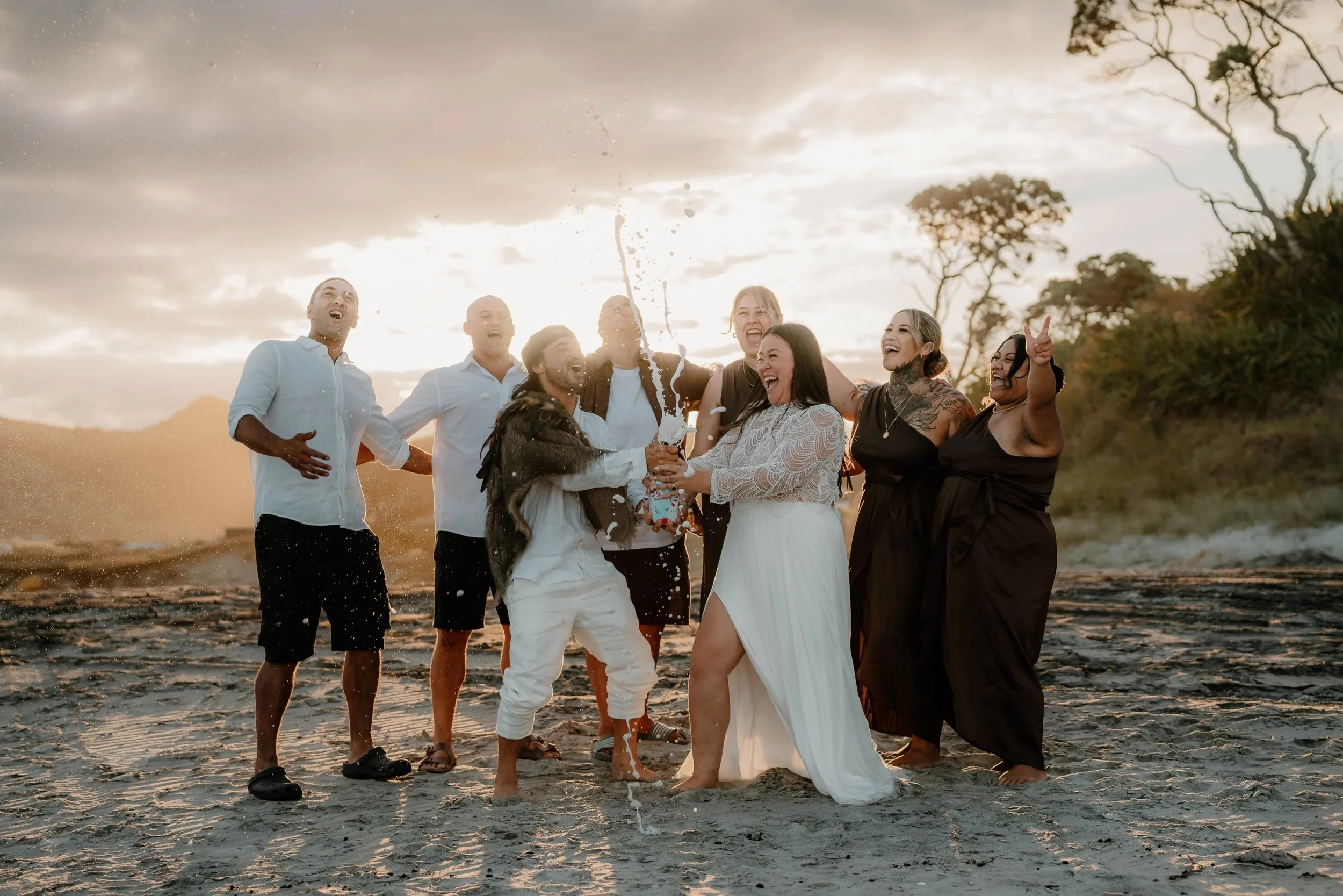 Vibrant beach wedding celebration at sunset, showing the couple and their bridal party laughing and spraying champagne in warm golden light.