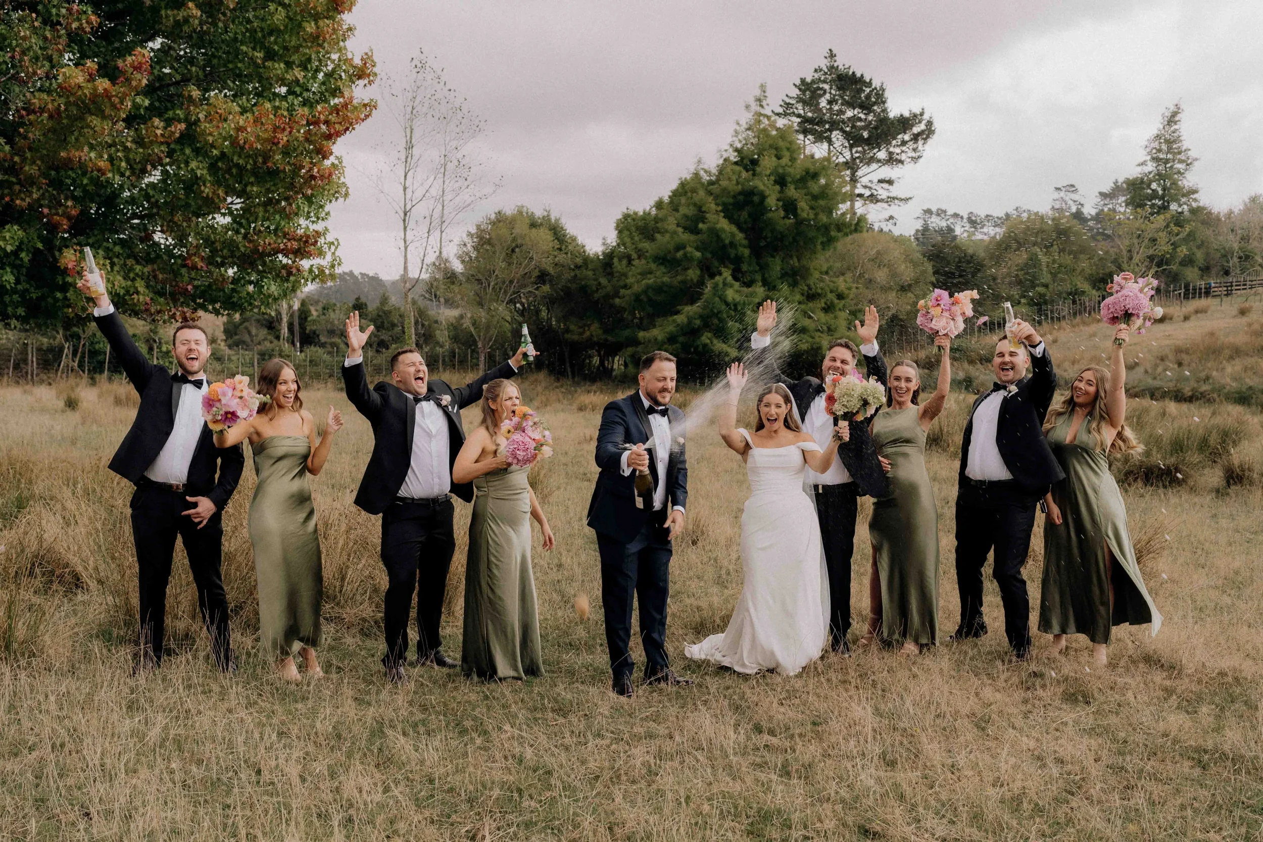 Vibrant outdoor wedding photo capturing the bridal party cheering as the groom opens champagne in a rustic field backdrop.