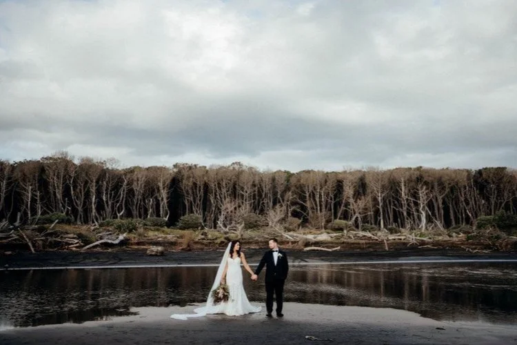 Moody coastal wedding portrait of a couple holding hands on a reflective shoreline, framed by driftwood and windswept forest.