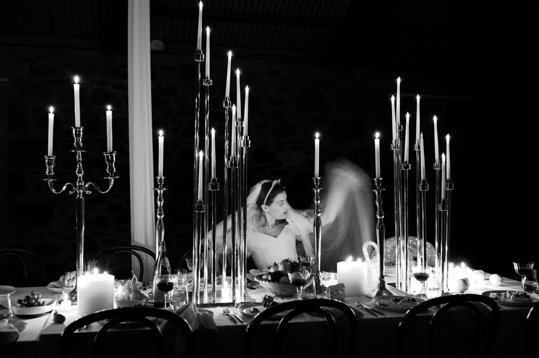 Dramatic black-and-white wedding photo of a bride at a candlelit reception table with tall taper candles, photographed in an editorial, fine-art style.