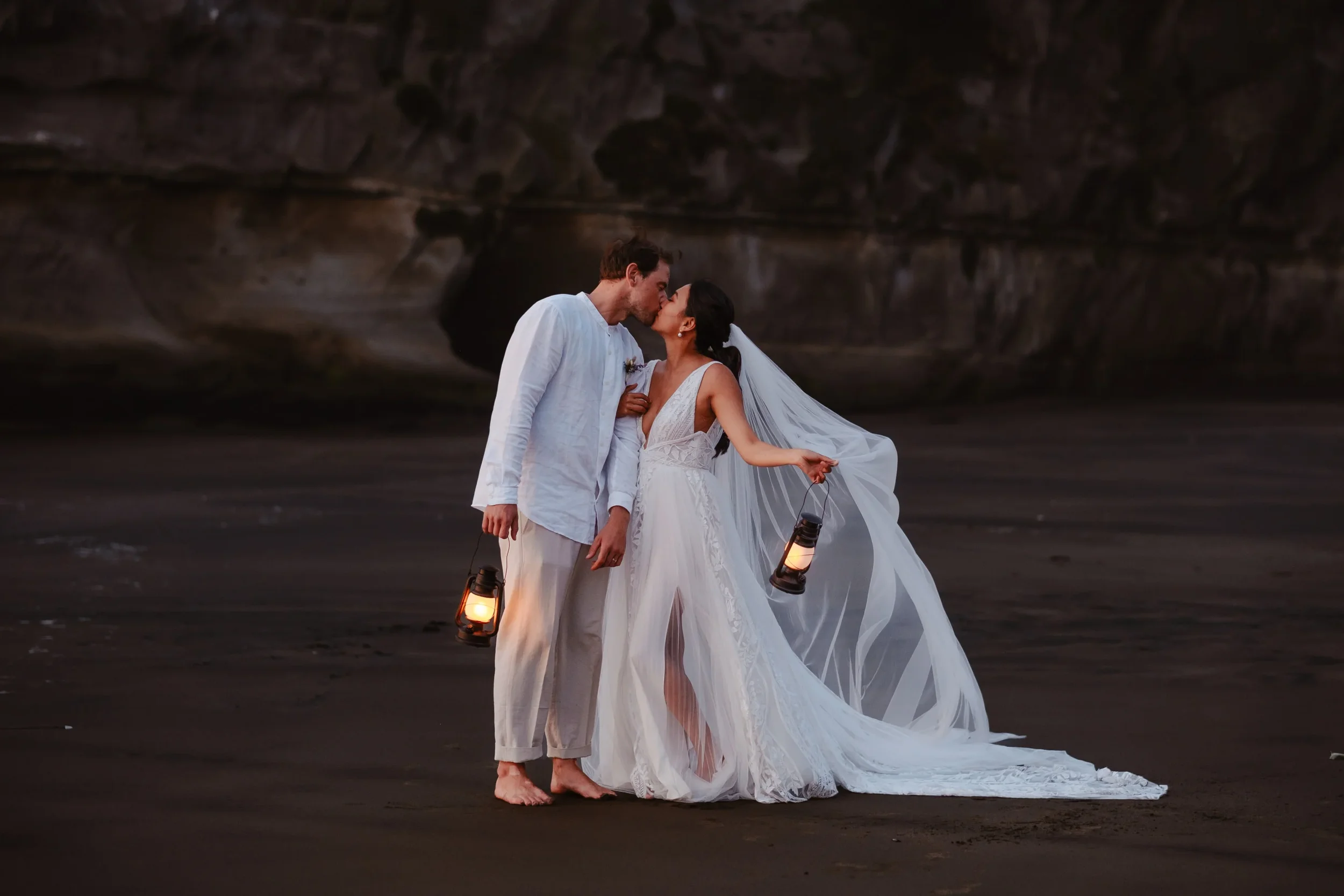 Moody black-sand beach wedding portrait of a bride and groom kissing at dusk while holding lanterns, photographed in a cinematic, atmospheric style.