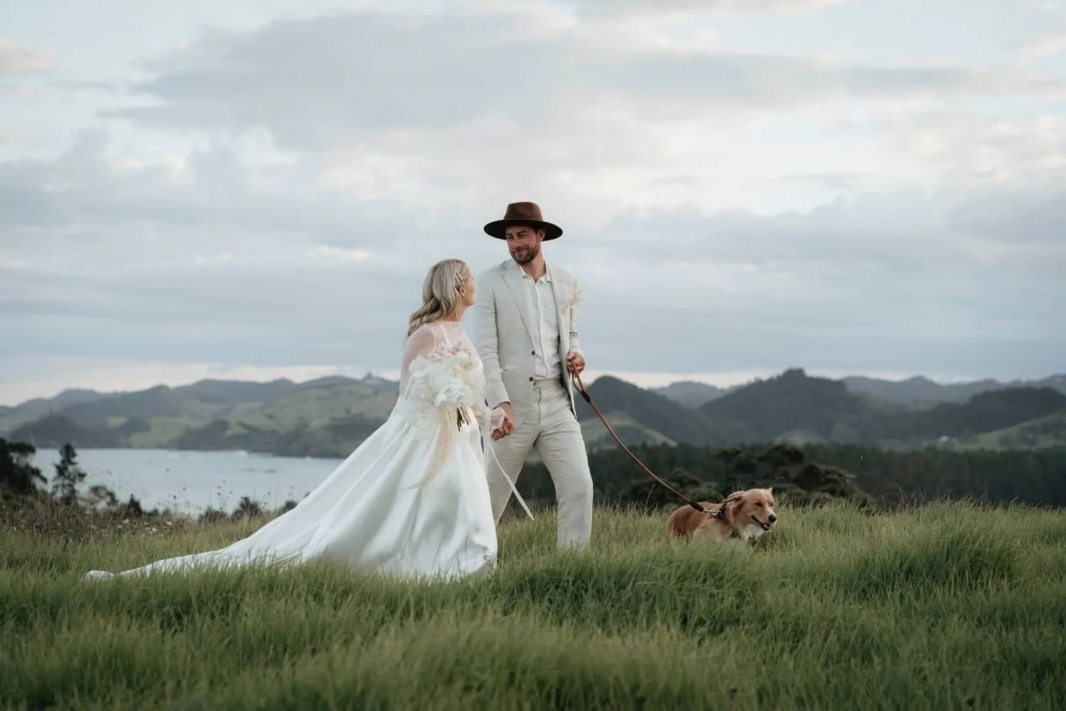 Elegant outdoor wedding portrait of a couple and their dog on a grassy hilltop with ocean views, showcasing natural, documentary-style photography.