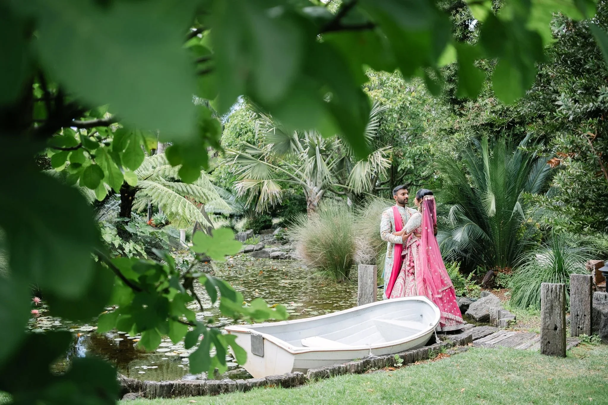 Colourful garden wedding portrait of a couple in traditional attire standing by a pond with a rowboat, captured through lush green foliage.