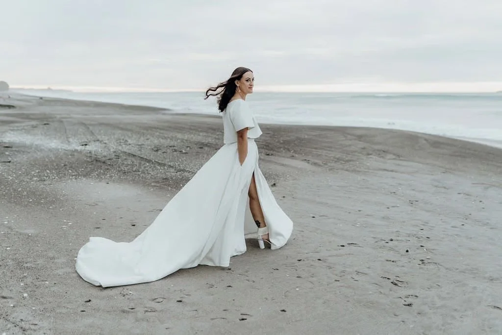 Beautiful beach wedding photo of a bride in a long, flowing gown walking beside the ocean, photographed in soft, natural light.