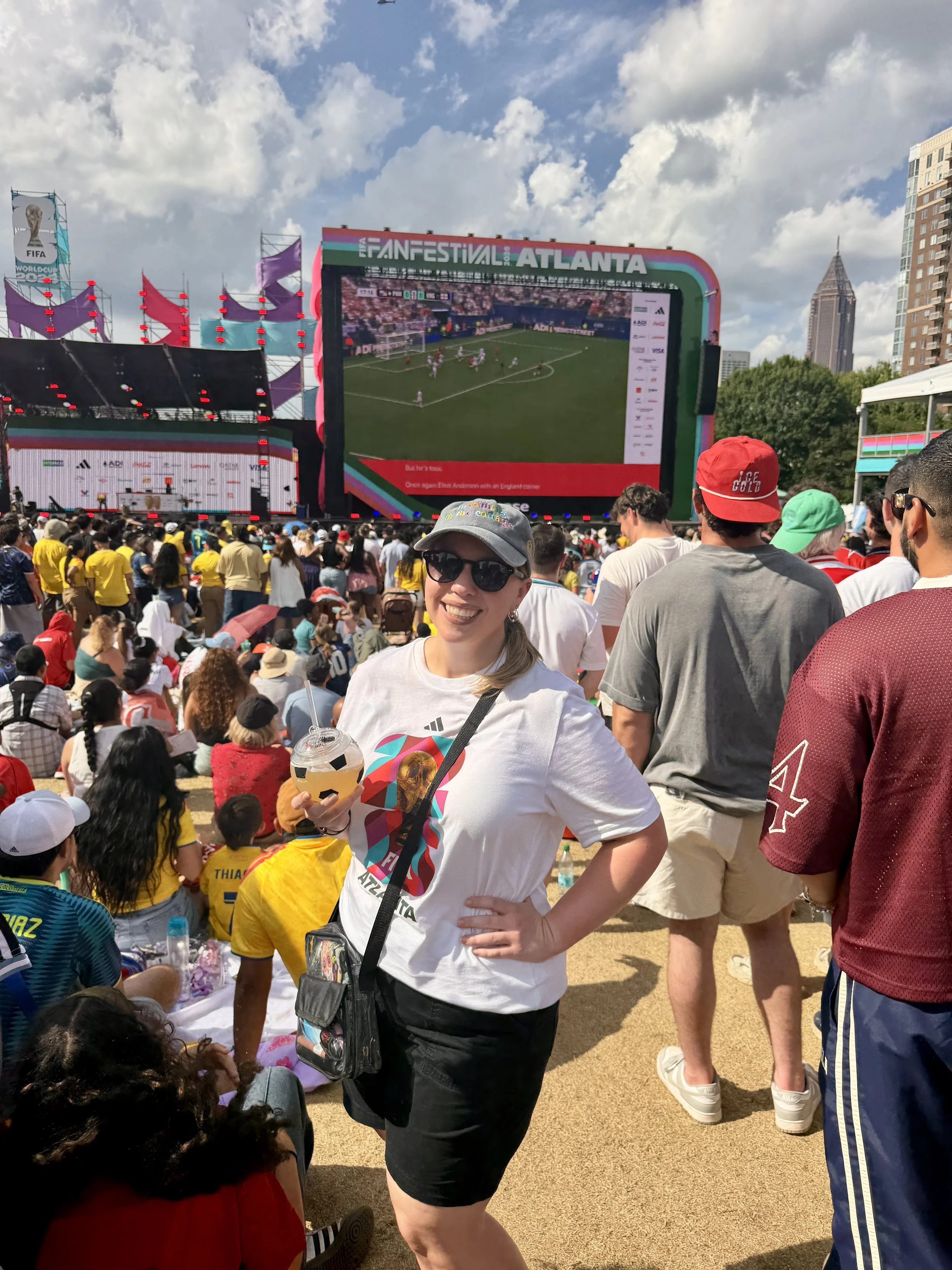 Meredith, LPC wearing a white FIFA Atlanta t-shirt, holding a soccer ball shaped cup, standing in front of a large screen showing a World Cup soccer game with a large crowd of fans and Atlanta buildings in the background